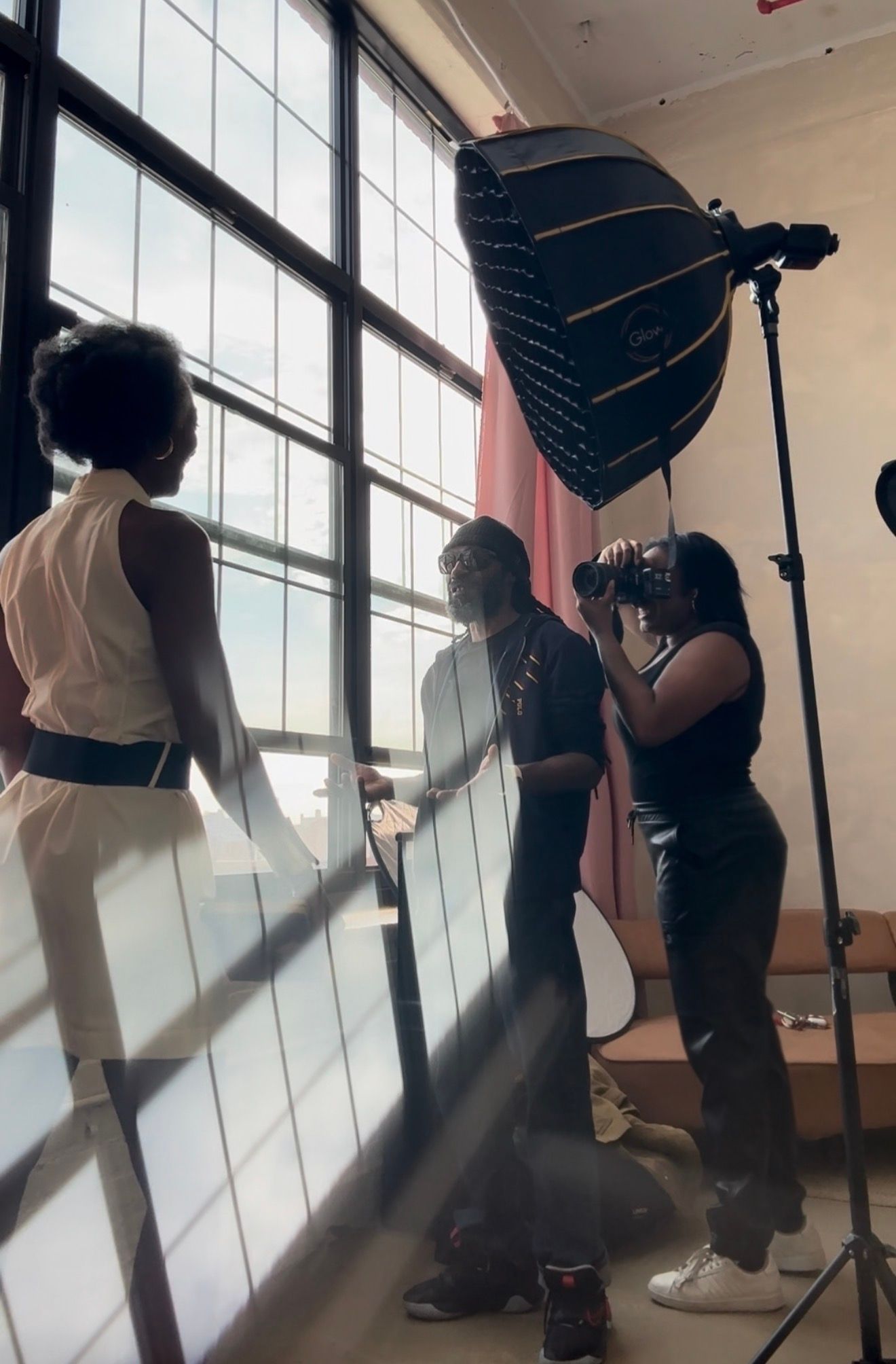 A group of people, including a woman in a white top, are setting up lighting equipment in a studio setting with large windows.