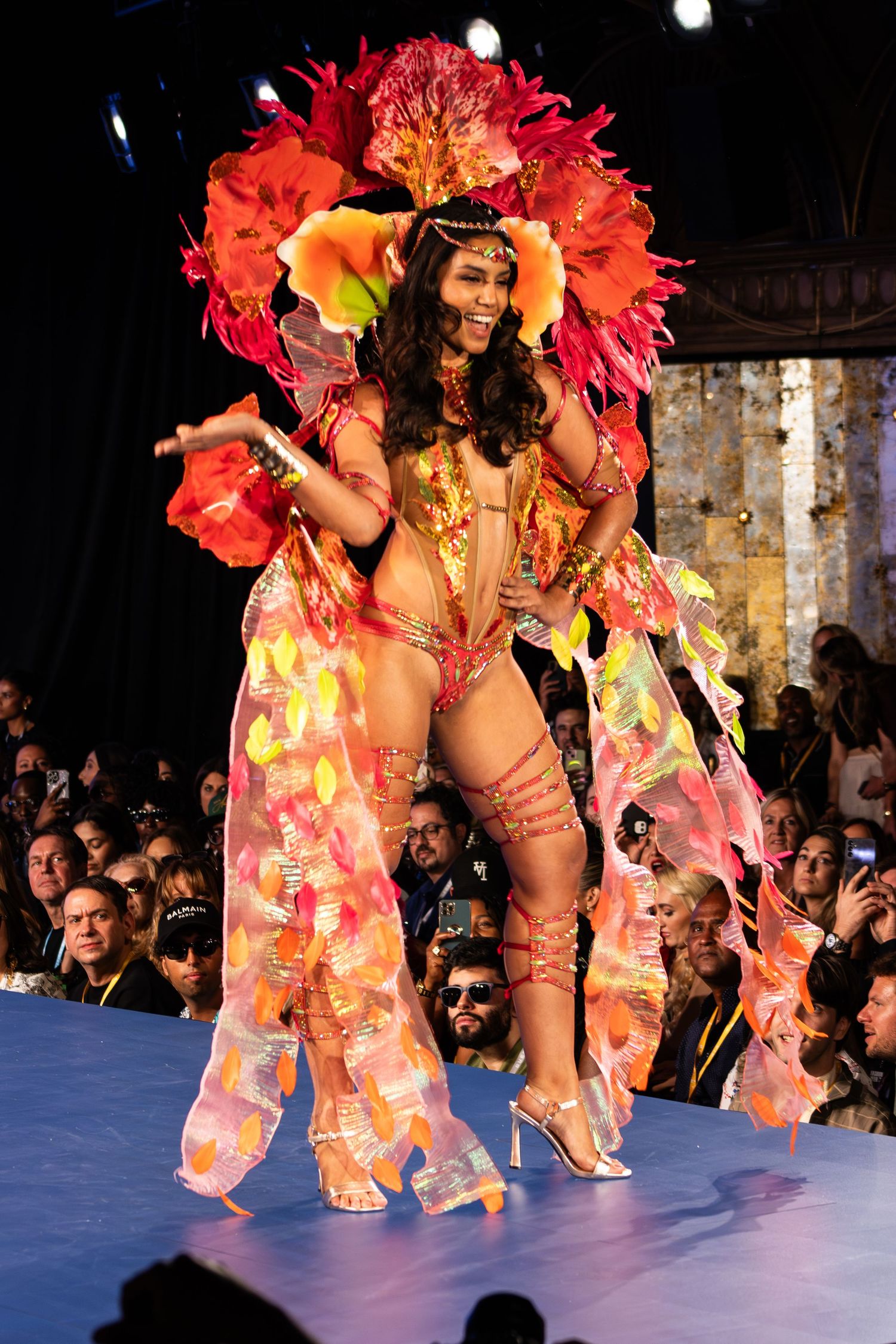 Model poses in stunning red and orange feathered carnival costume with large floral headpiece on runway.