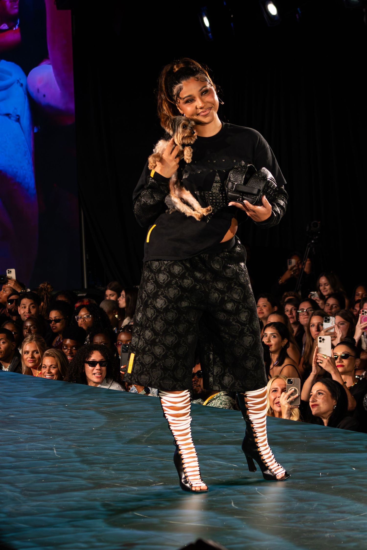 Model carries small dog on runway wearing black patterned culottes with strappy heeled boots at fashion show.