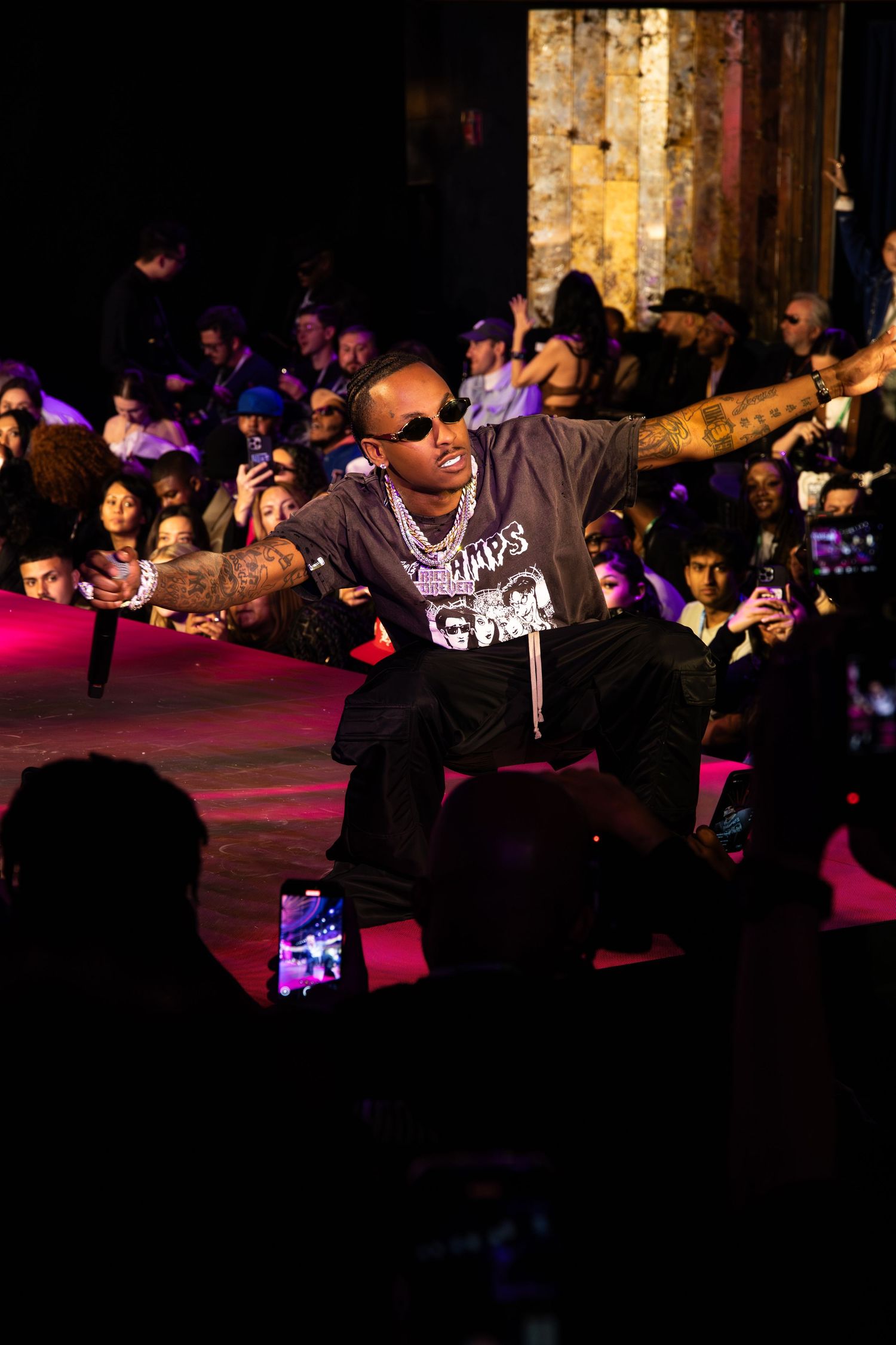 Performer in sunglasses and dark clothing crouches on stage during a nightclub performance with purple lighting and crowd.