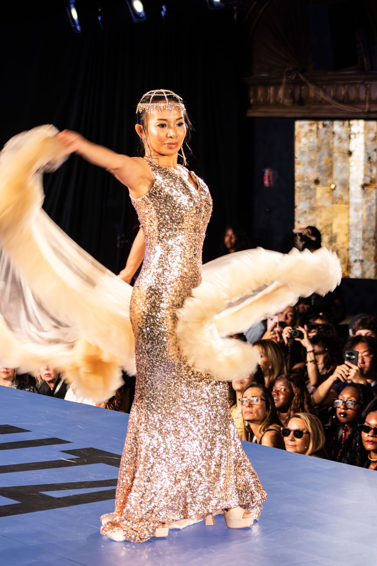 Model in gold sequin gown with feather wrap poses on fashion show runway.