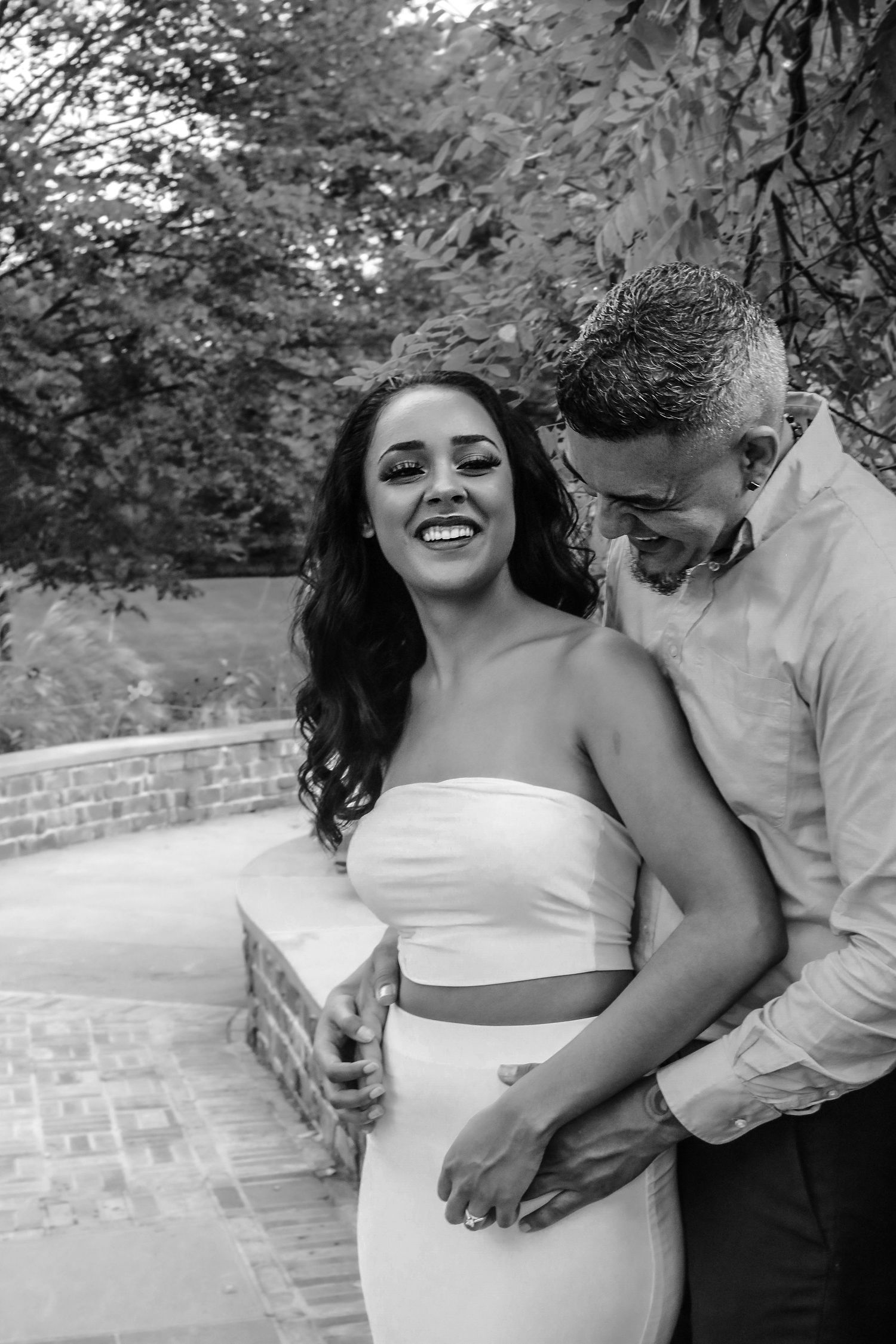 A romantic black and white photo series shows a couple embracing and laughing together on a brick wall outdoors.