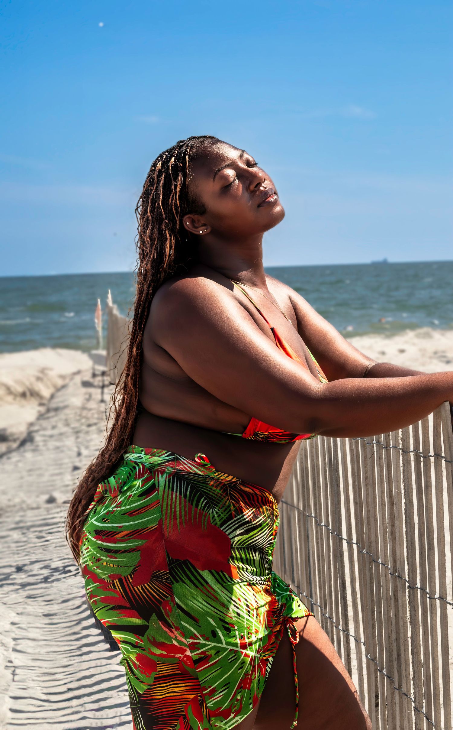 Person in tropical print sarong enjoying a sunny day leaning against white fence at beach with ocean view.