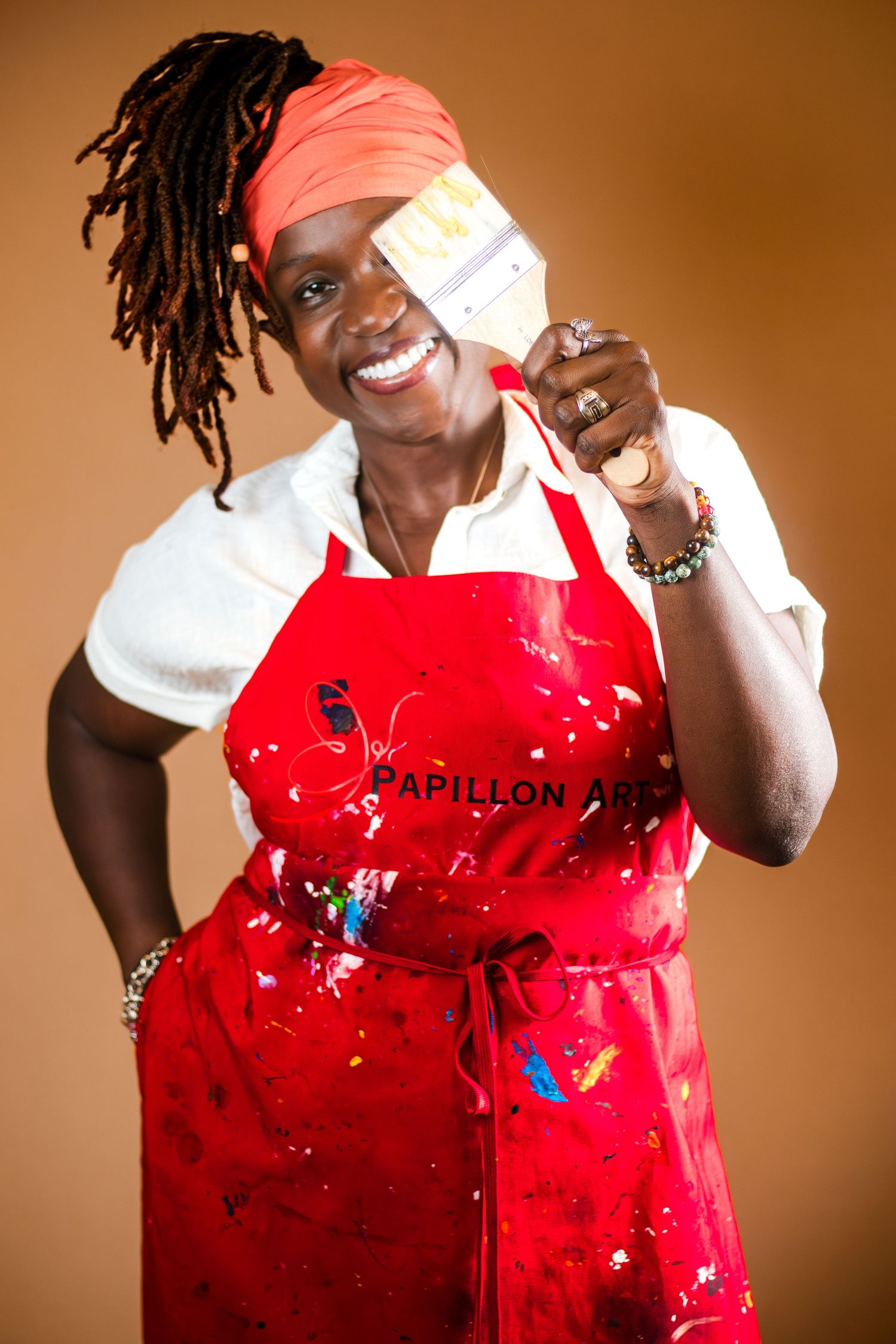 A chef wearing a red apron and headscarf smiles while holding a paintbrush in a professional portrait against brown background.