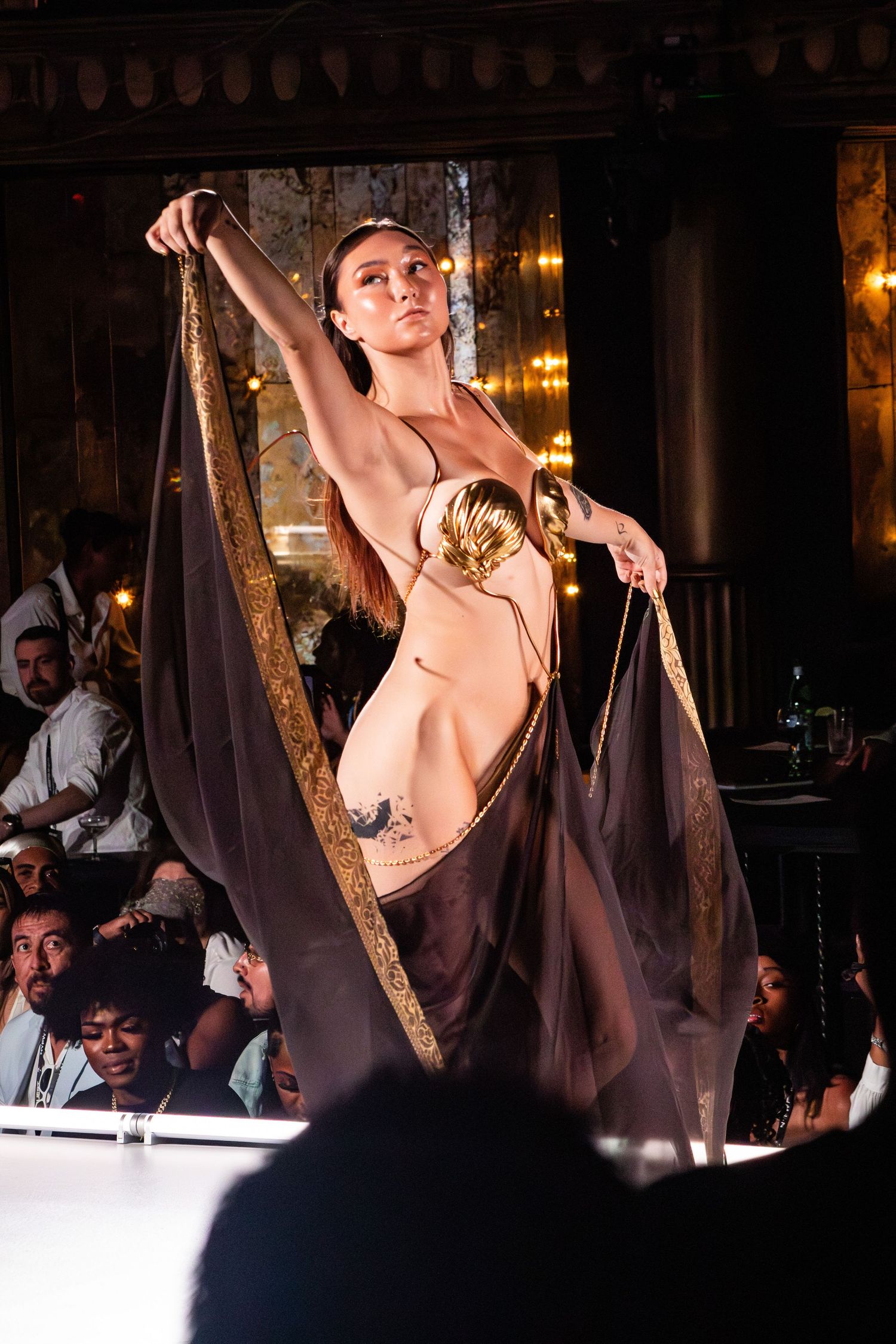 Performer in nude-colored costume strikes elegant pose on stage with dramatic lighting.