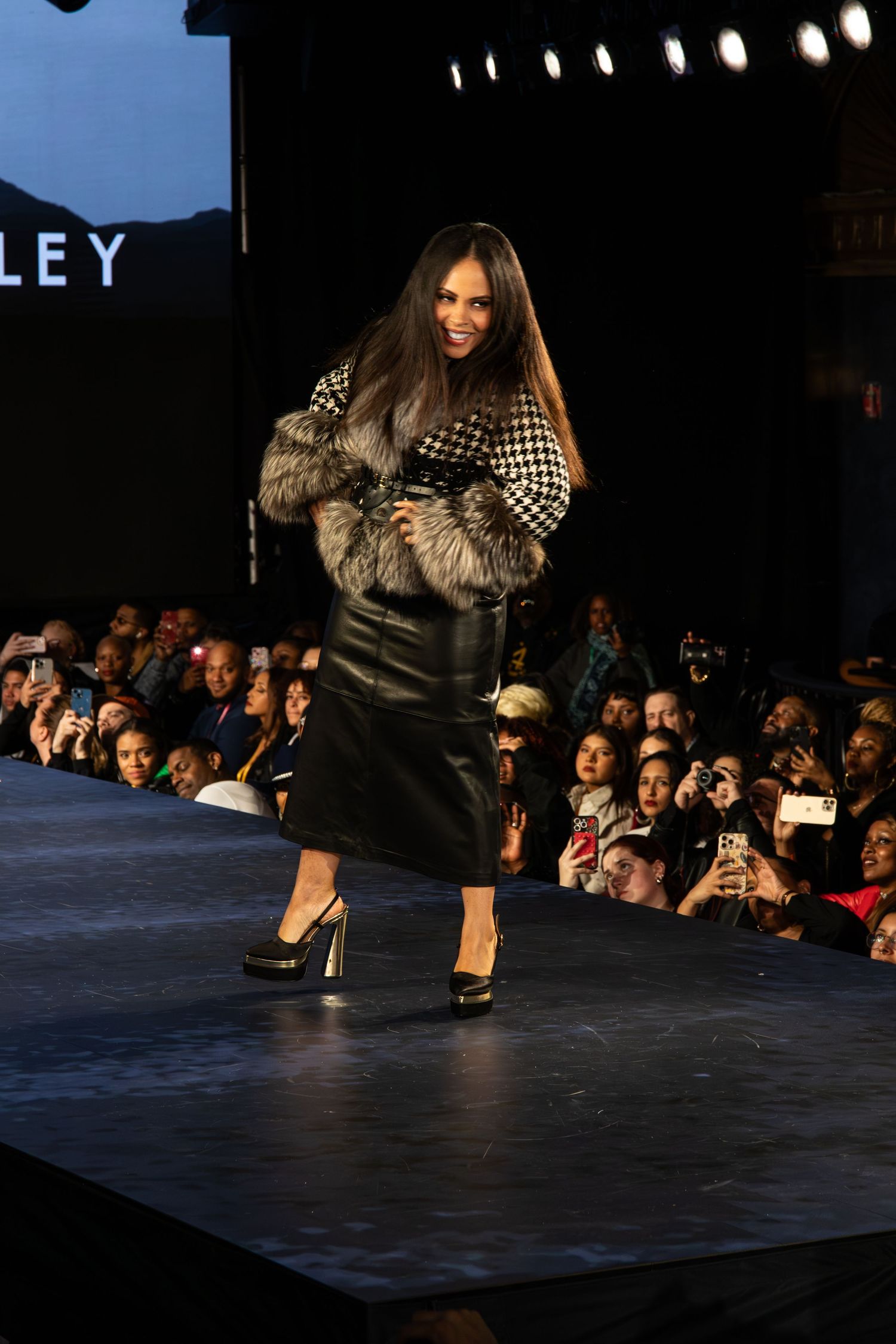 A fashion model walks down a runway wearing a black leather skirt, fur wrap and checkered top at a fashion show.