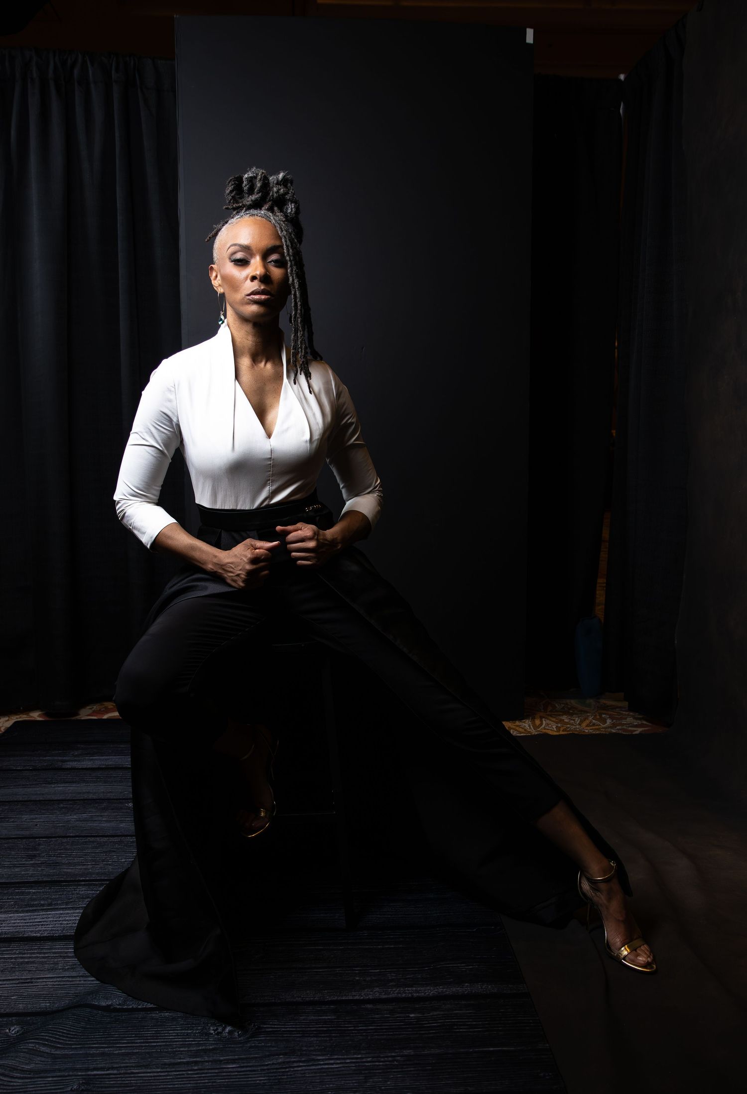 Elegant woman in white blouse and black wide-leg pants sits confidently against dark backdrop with braided updo.