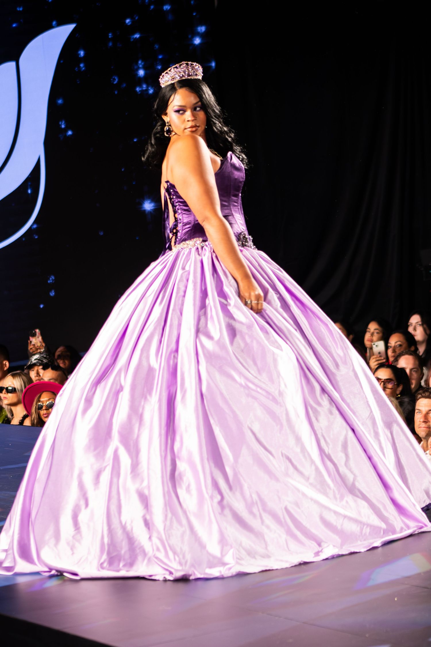 Model in lavender ball gown with tiara walks fashion show runway.