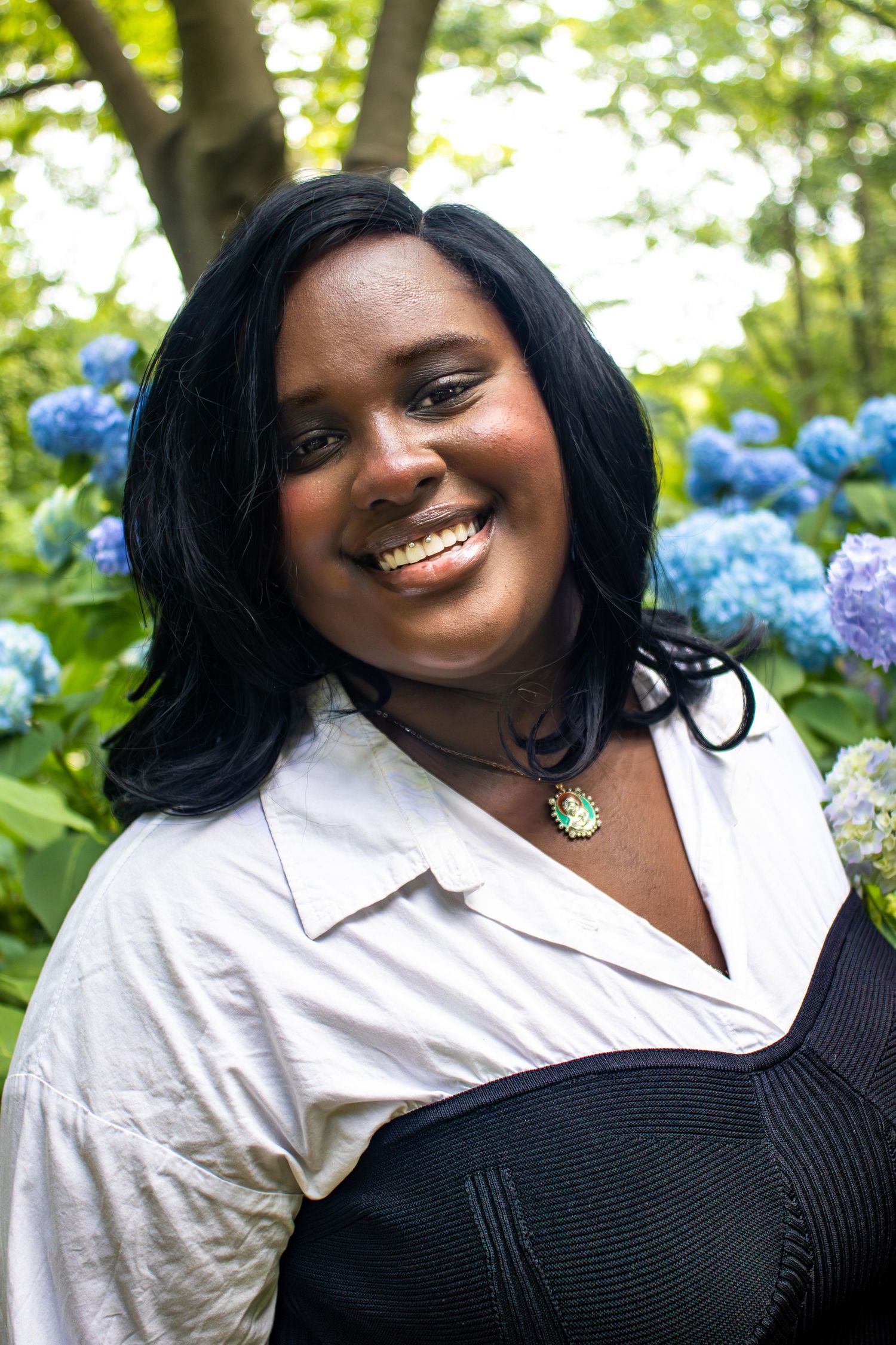 A radiant smile against a backdrop of blue hydrangeas in a sun-dappled garden setting.