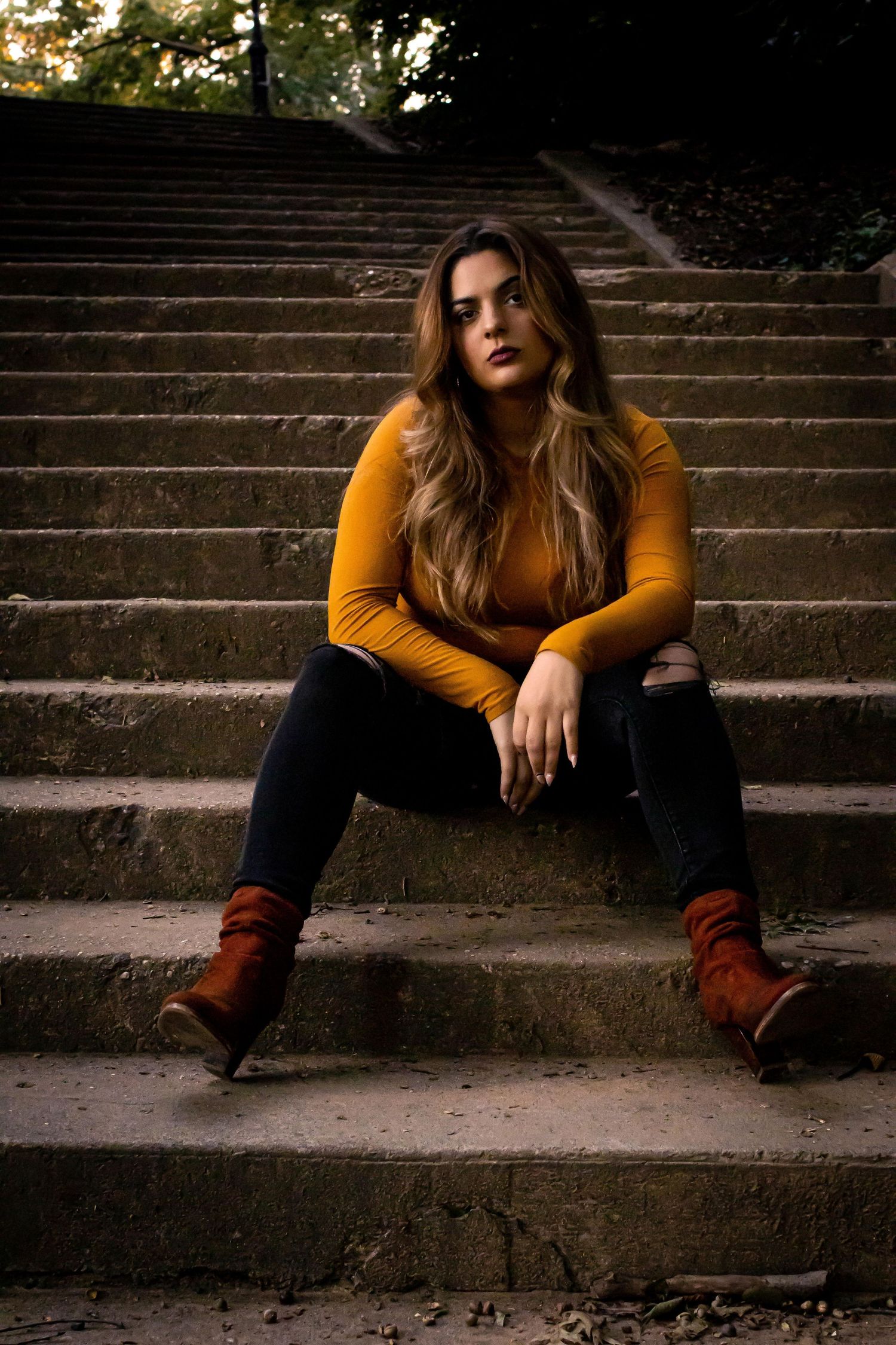 Person in mustard sweater and ripped jeans poses on concrete steps at dusk.