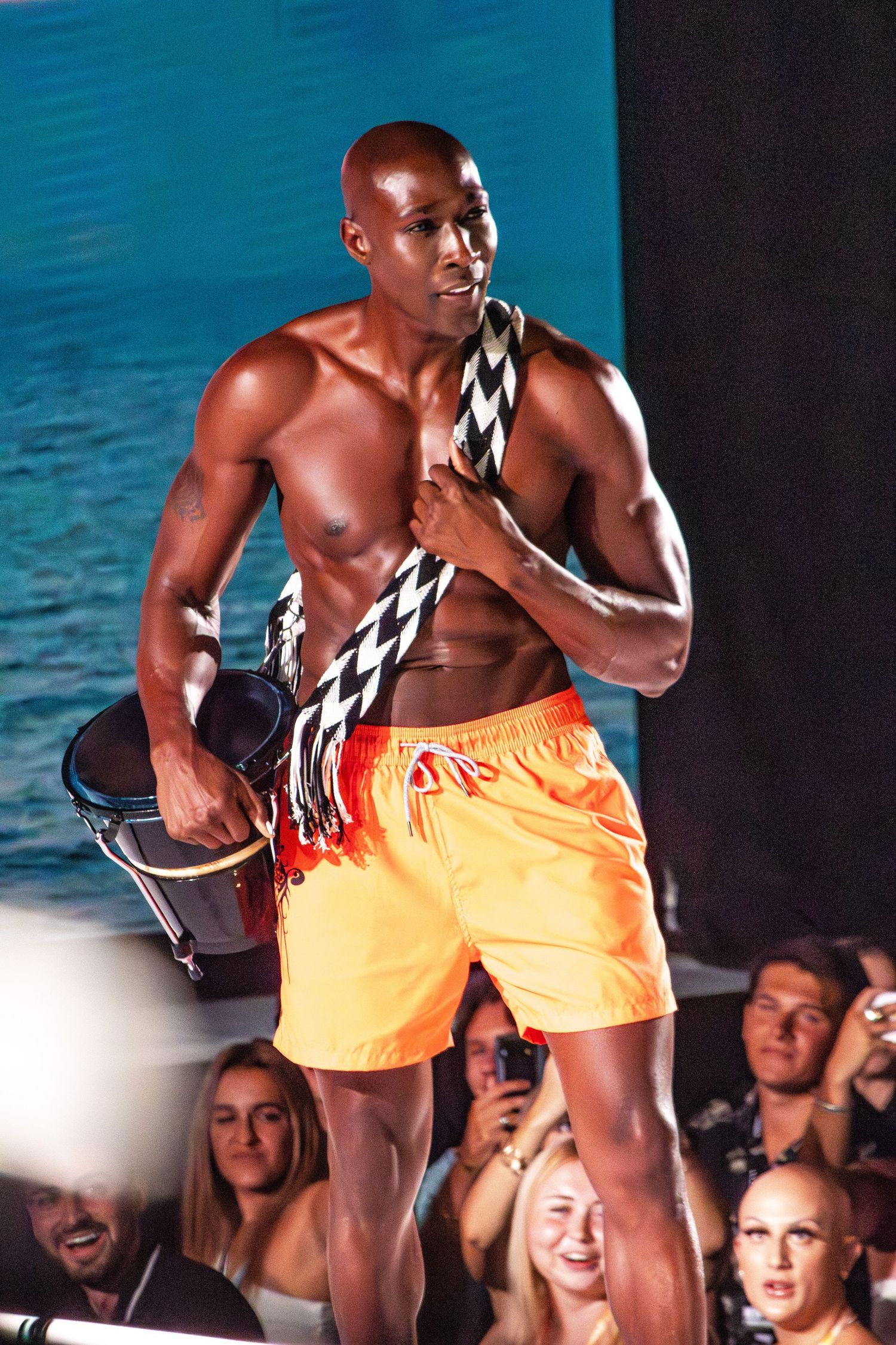 Model presents orange swim shorts and black geometric print accessory on beachwear fashion runway.