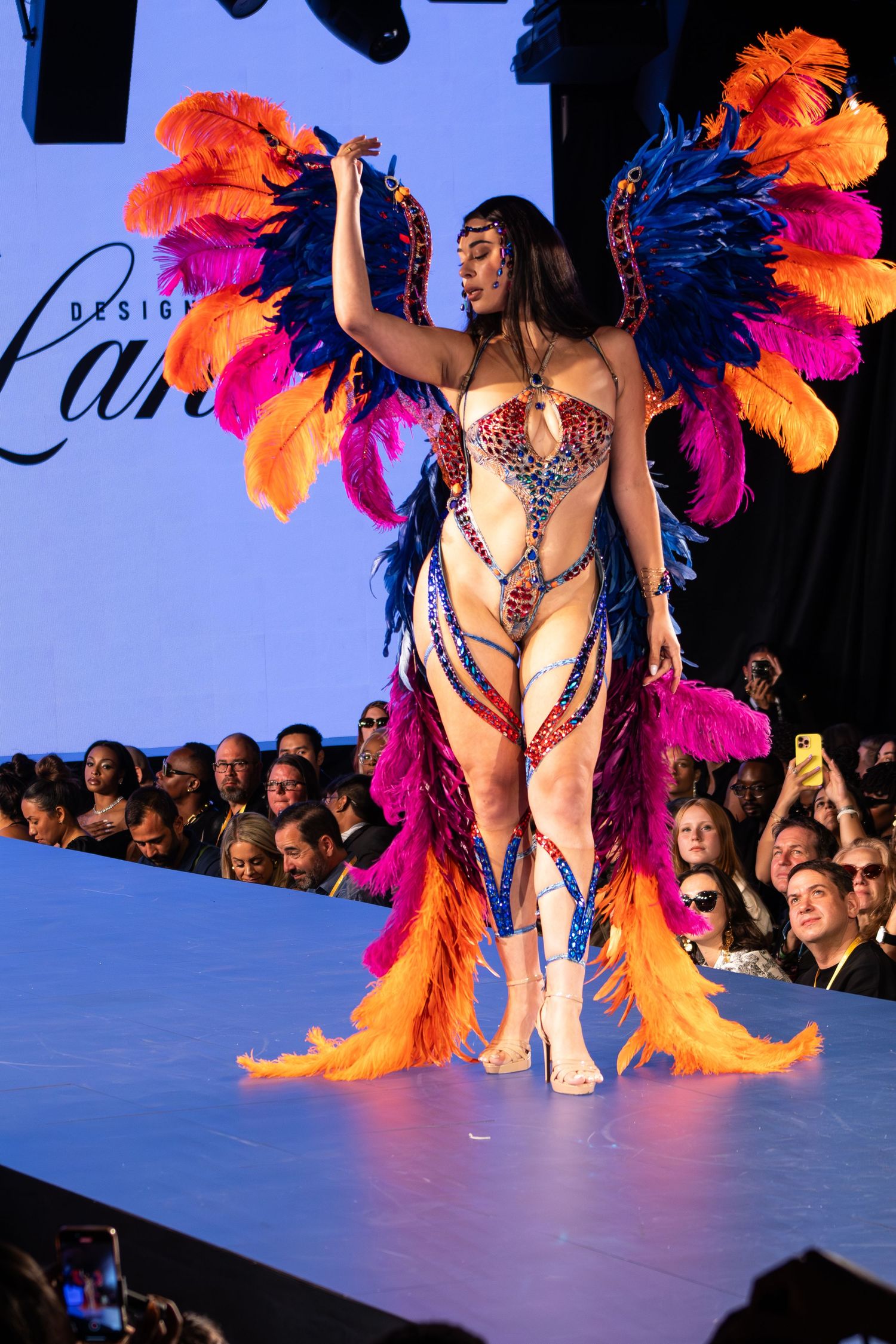 Model showcases vibrant carnival costume with colorful feather wings and jeweled bodysuit on fashion runway.
