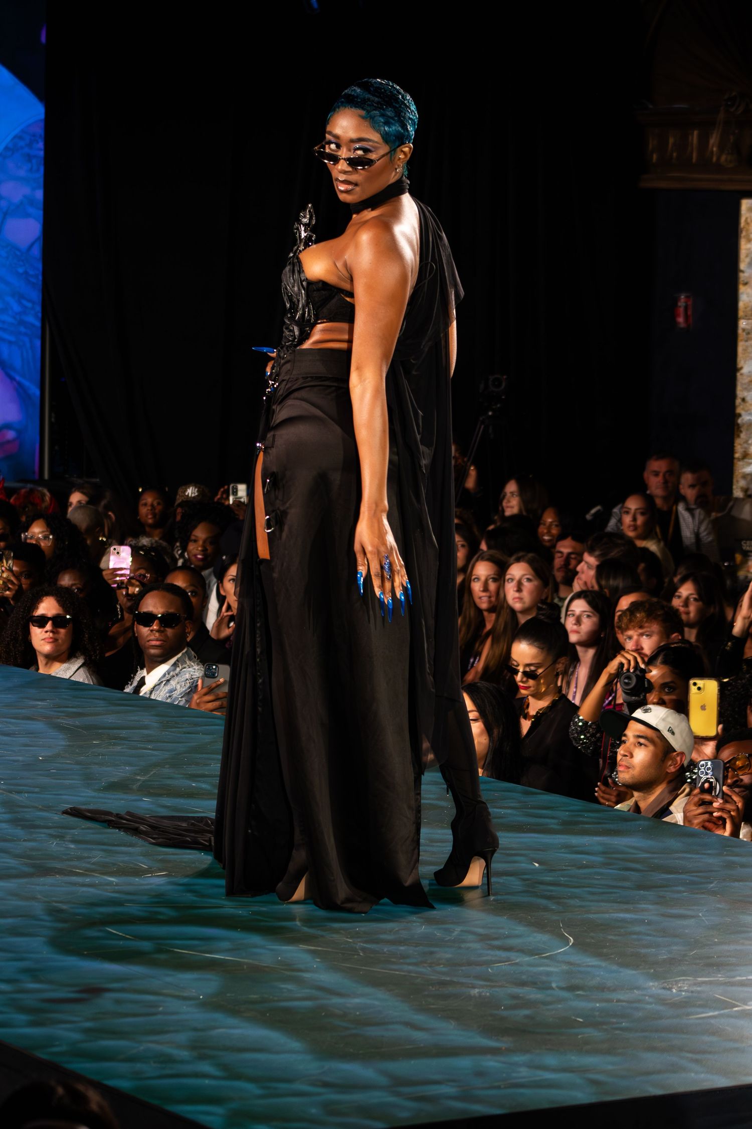 Model in black two-piece outfit with flowing skirt and blue hair walks runway at fashion show.
