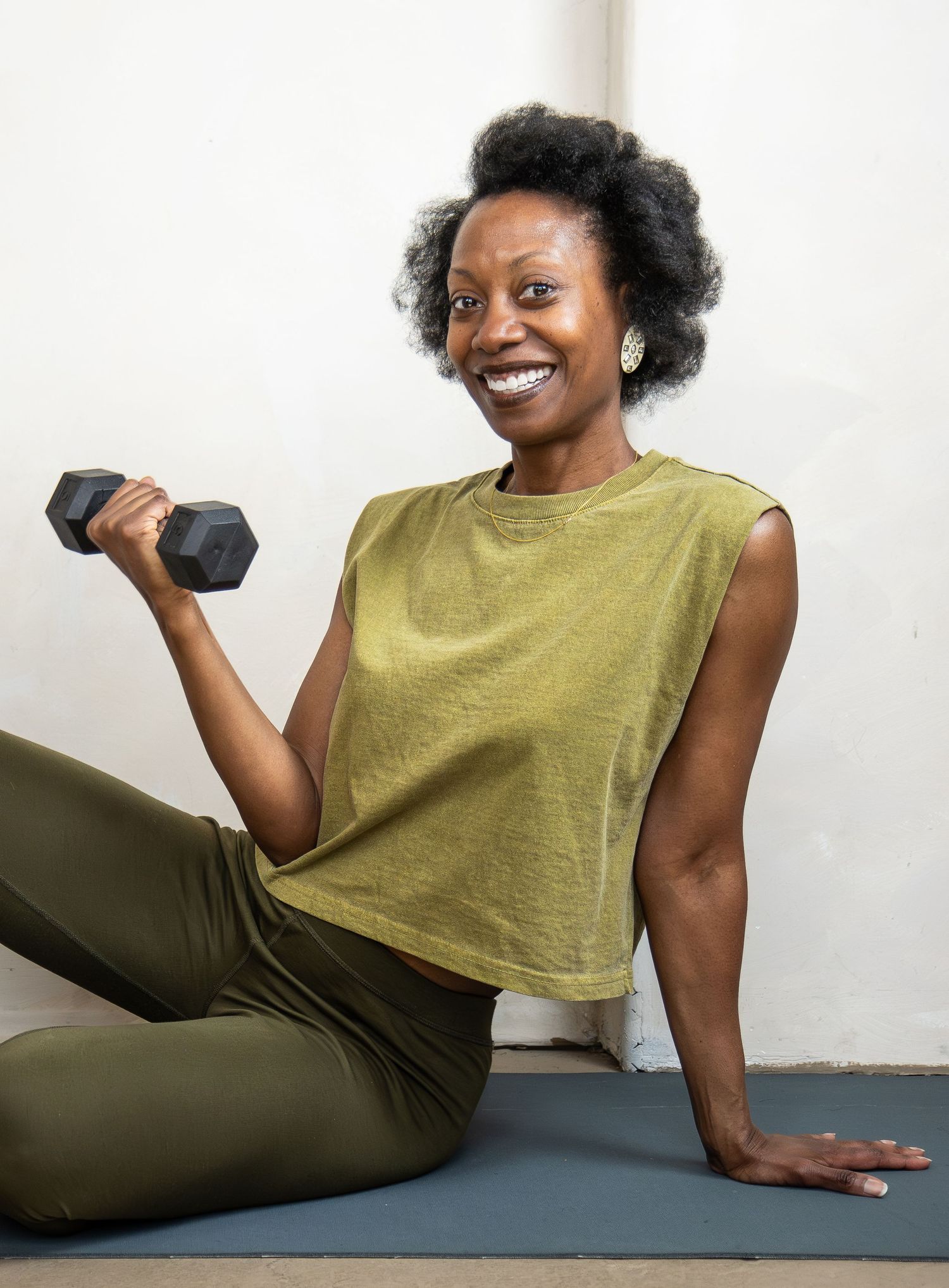 A smiling person in olive workout attire sits on a yoga mat while holding a dumbbell during an exercise session.