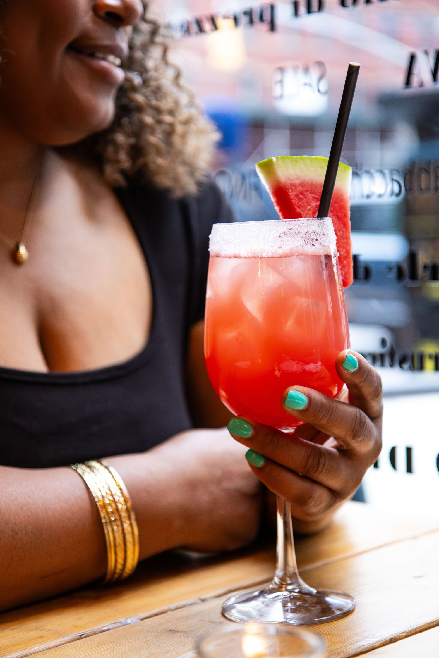 Woman with green nails holding a watermelon cocktail in a stemmed glass with a black straw at a bar.