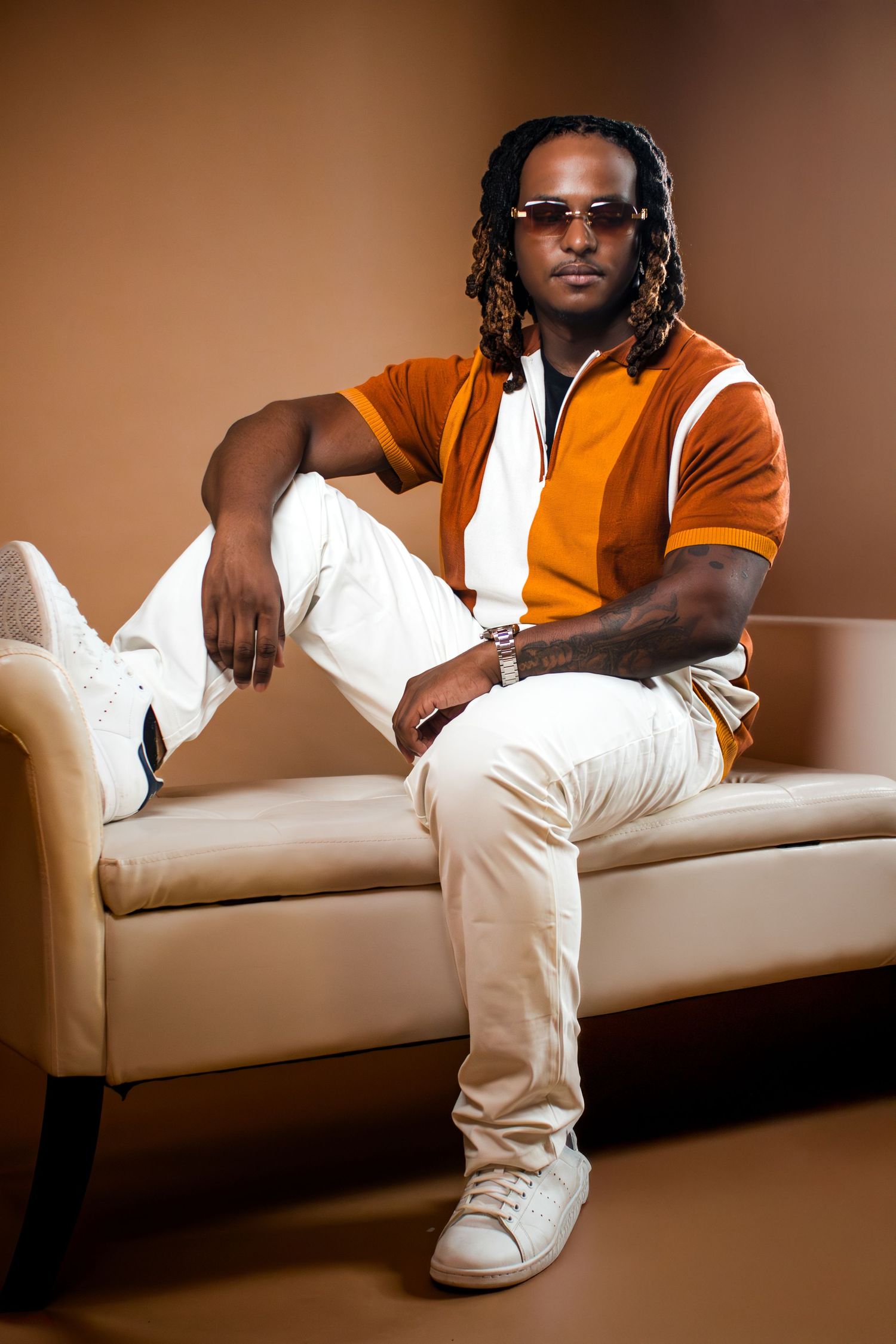 Stylish person in orange polo shirt and white pants relaxing on beige couch wearing sunglasses and jewelry.