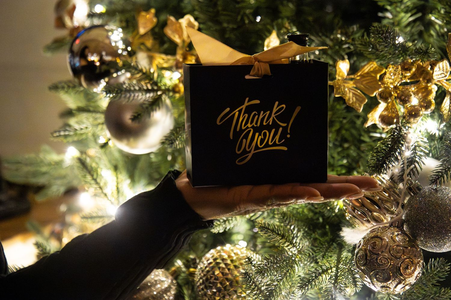 Hands holding a black Thank You gift bag with gold ribbon in front of a decorated Christmas tree with lights.