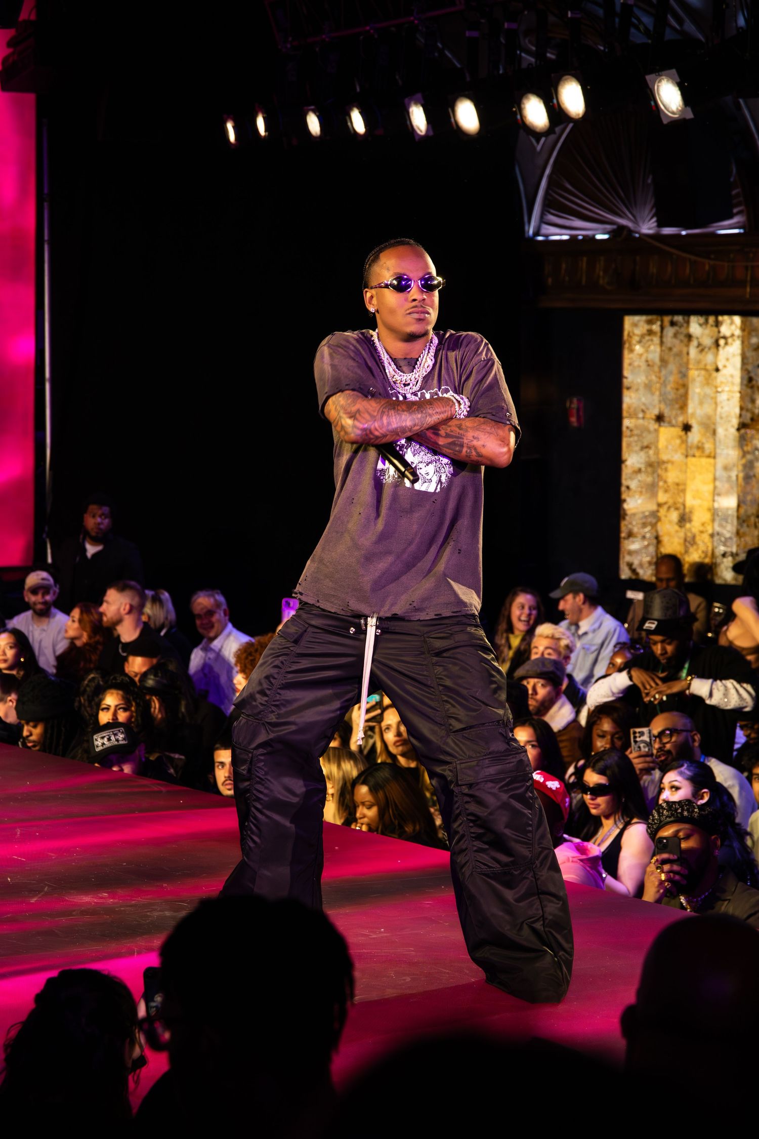 Performer in sunglasses and baggy pants energetically dances on a stage lit with dramatic purple lighting during a live show.