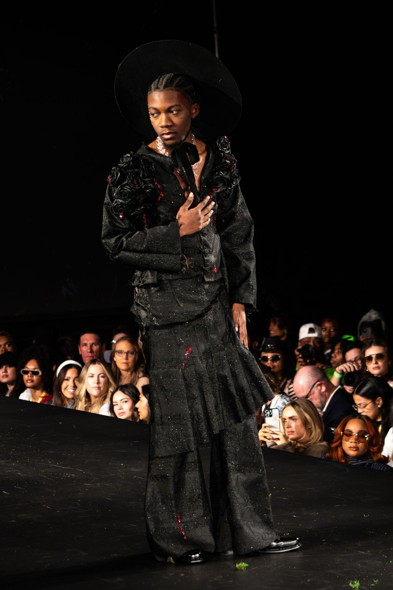Model walks down dark runway wearing all-black ensemble with wide-leg pants and fitted jacket at fashion show.