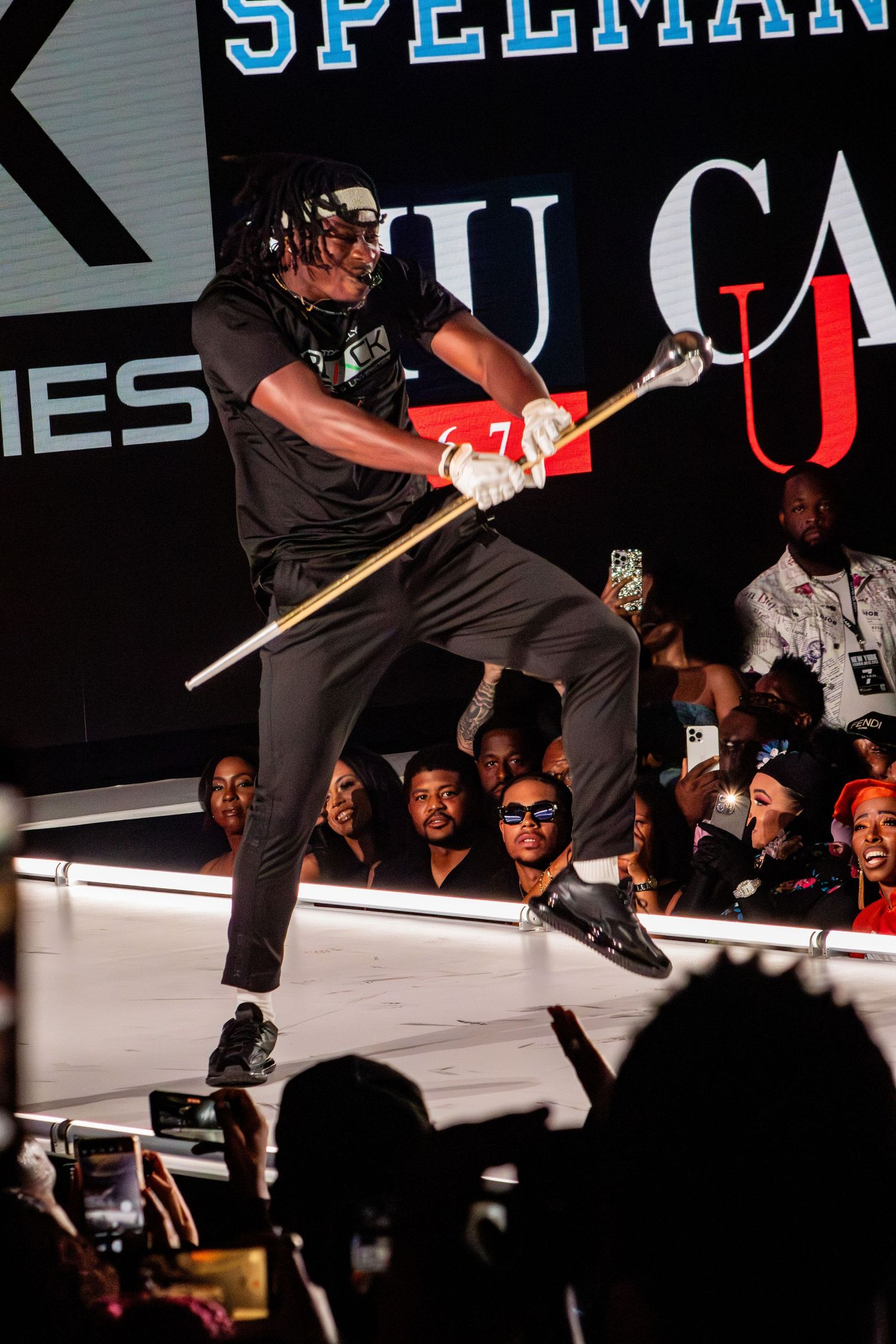 Energetic stage performer leaps while wielding a prop staff against lit backdrop.
