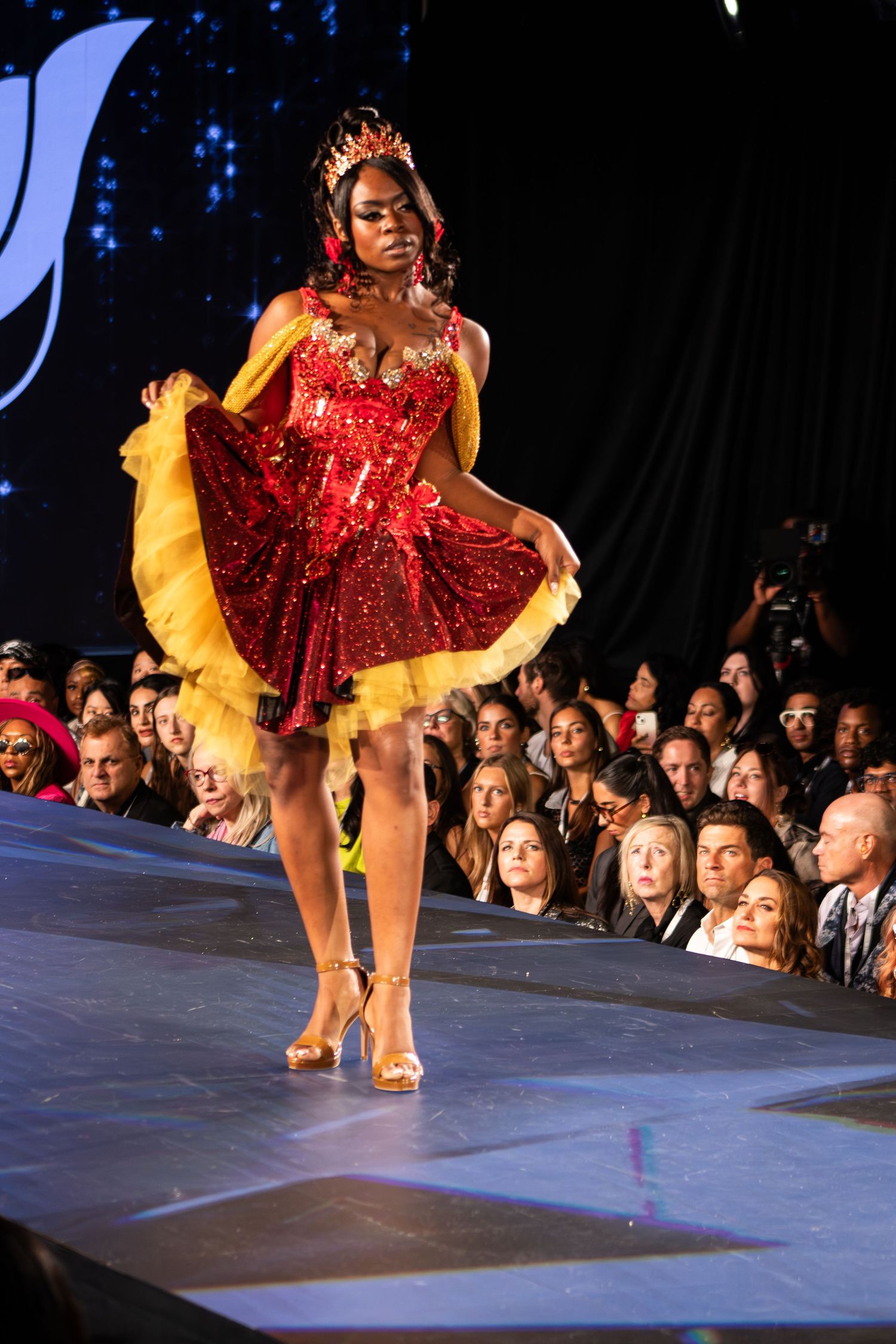 Model in red sequin dress with yellow tulle and crown walks fashion runway.