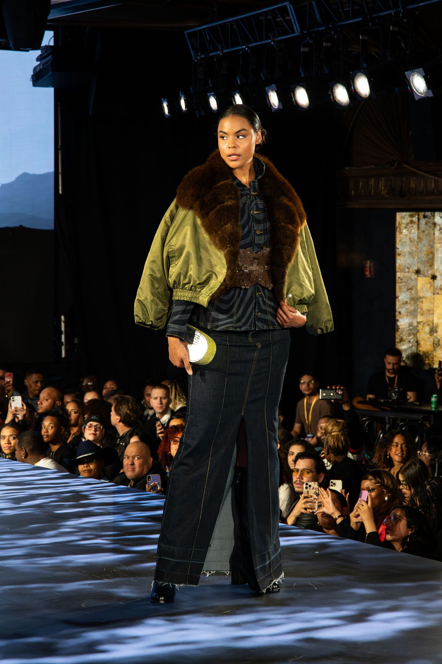 Model walks runway in metallic gold bomber jacket with fur collar over dark denim at fashion show with stage lighting.