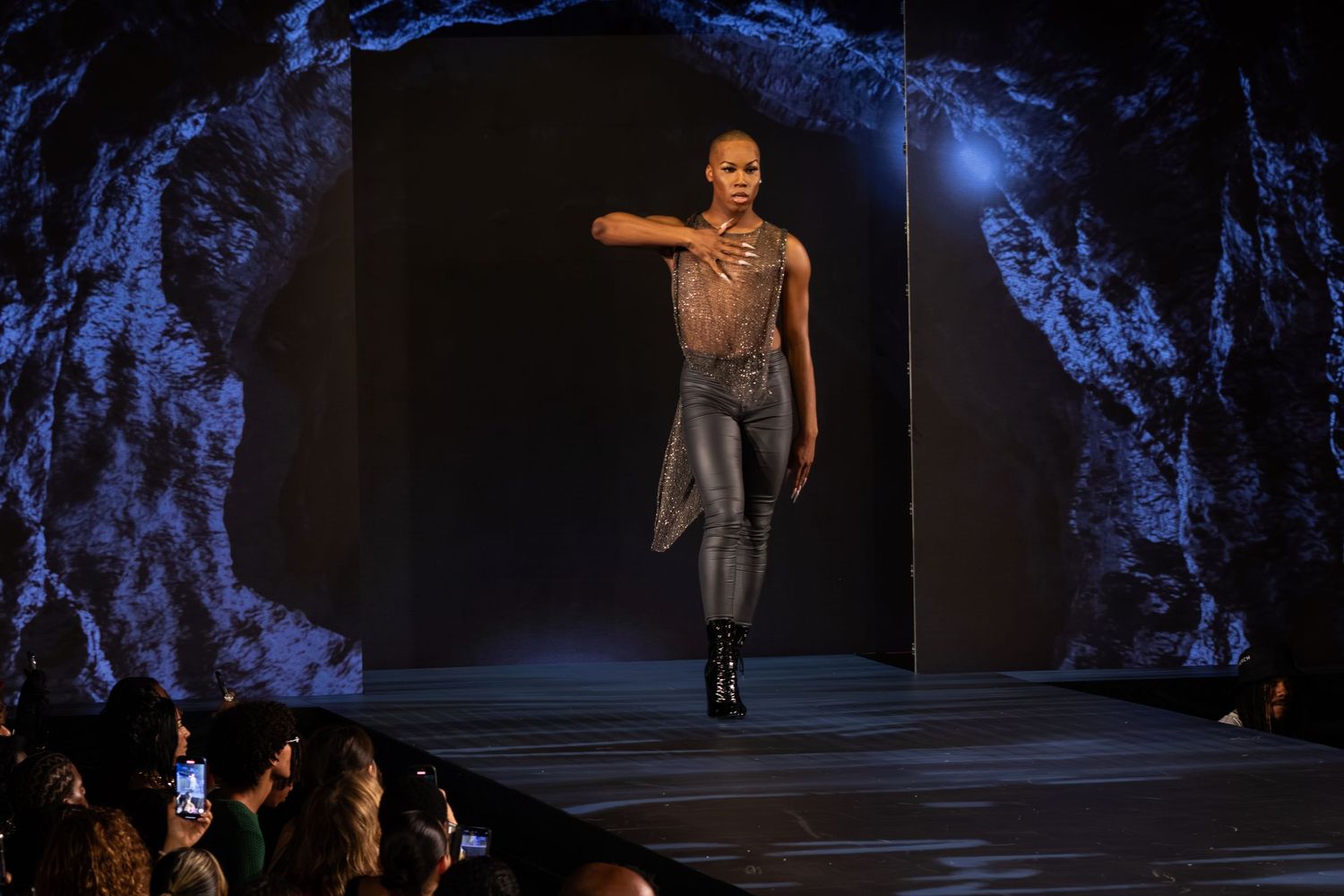 Female model poses on runway in metallic mesh bodysuit and dark leggings with black boots at fashion show.