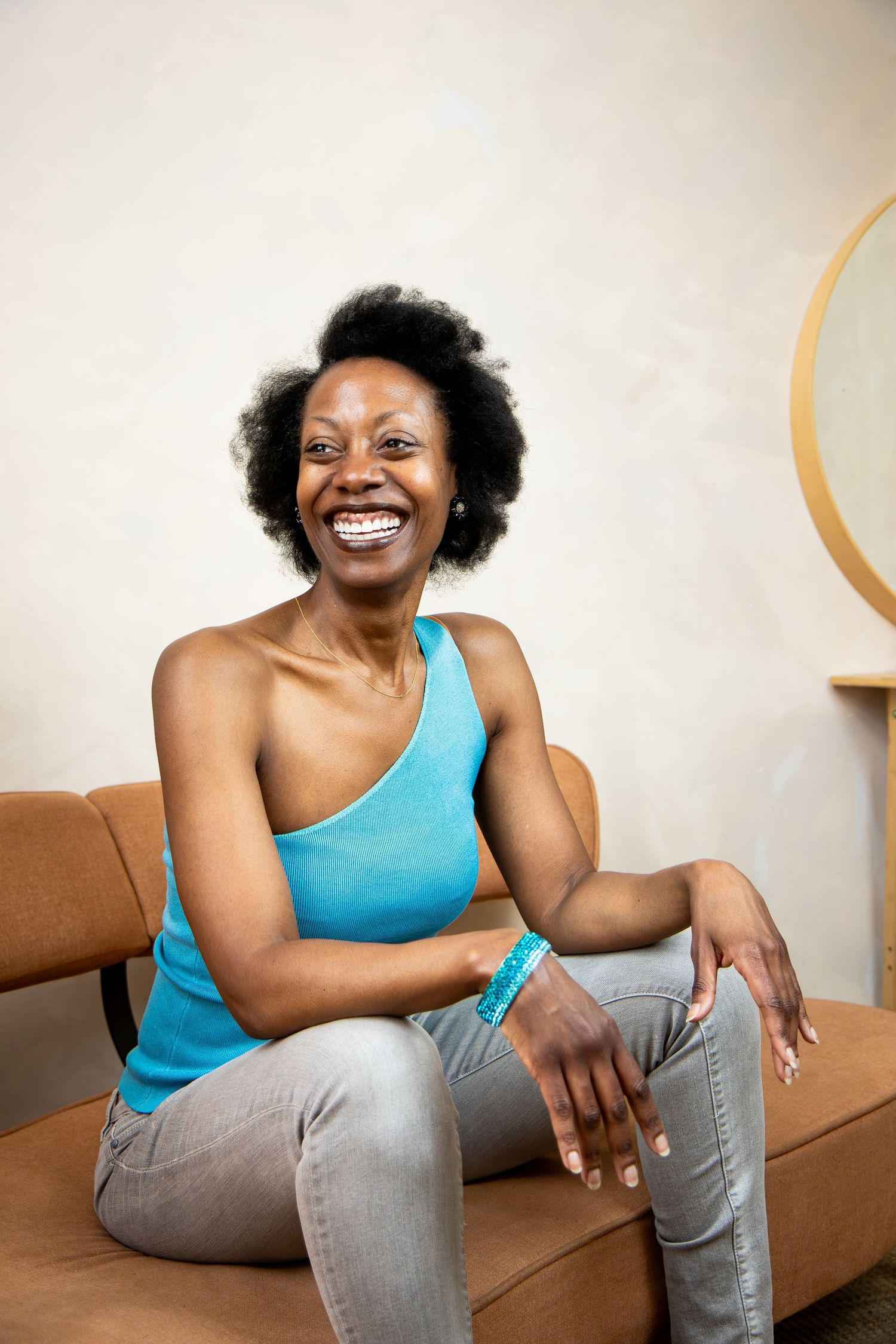 A joyful person in a blue one-shoulder top and gray jeans sits casually on a brown couch against a beige wall.
