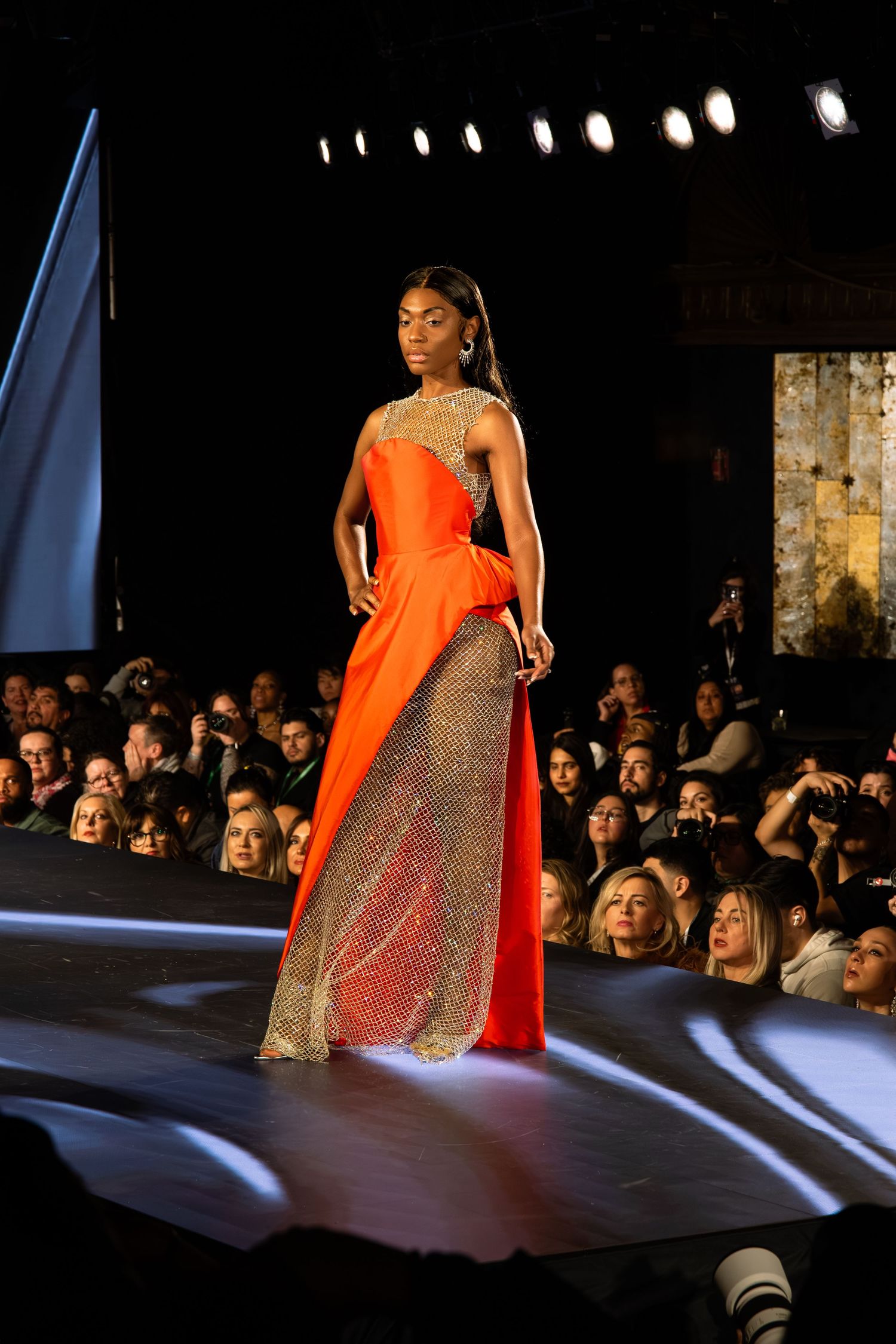 Model walks the runway in a stunning orange gown with glittering panels at a fashion show before a crowded audience.