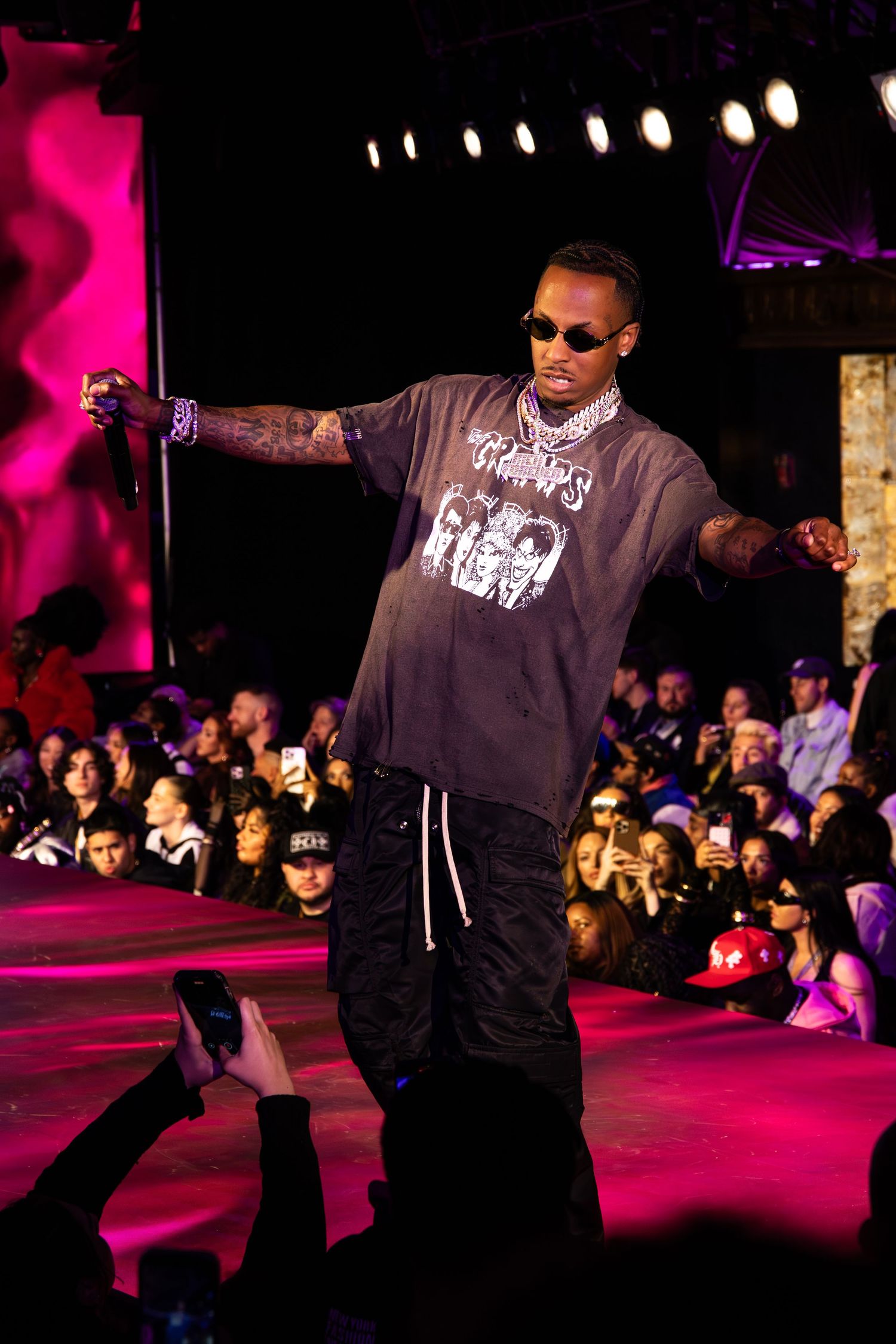 Performer in sunglasses and grey sweatshirt on stage with purple lighting and crowd visible in background.