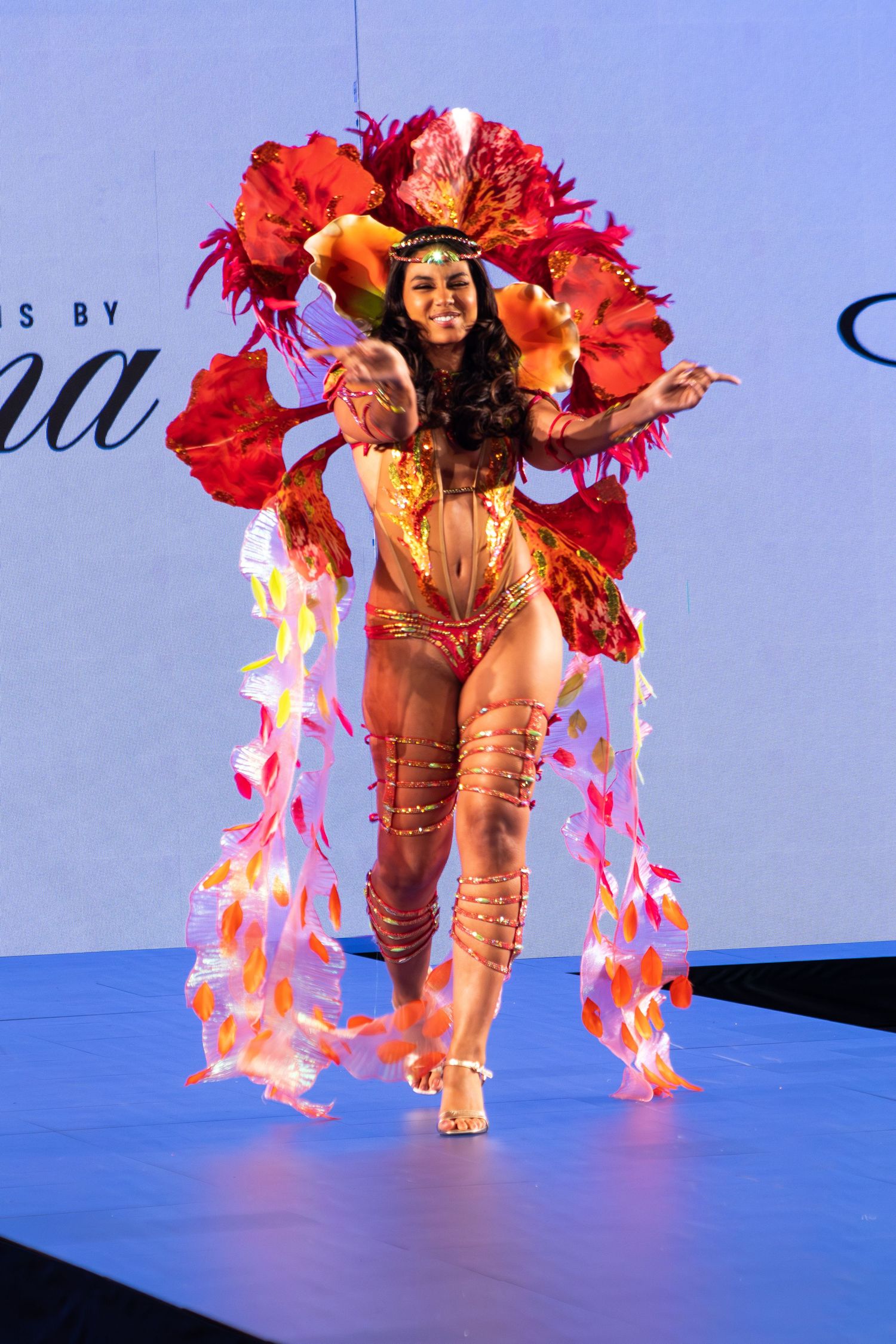 Model performs on runway in elaborate red and gold carnival costume with large floral headdress.