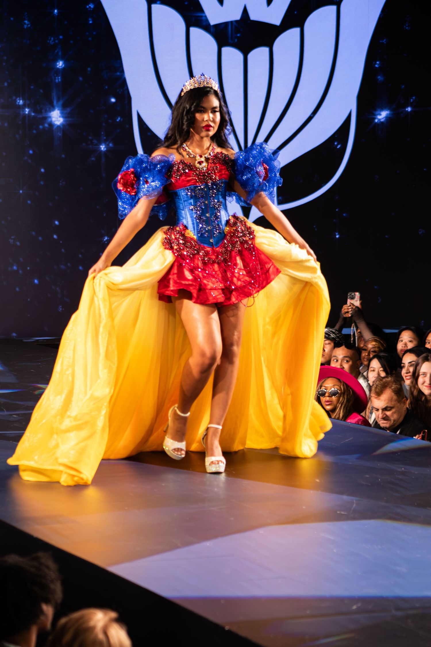 Model in Snow White costume with yellow skirt and tiara walks fashion show runway.