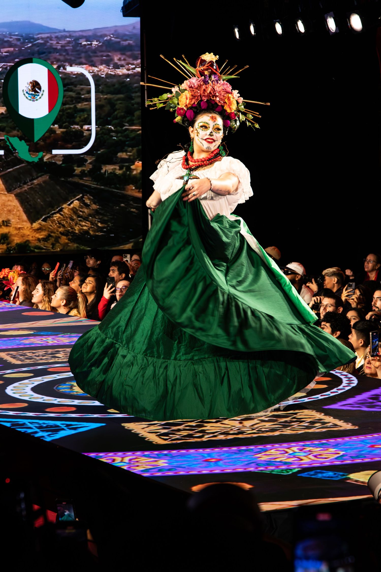 Traditional Mexican dancer performs in a flowing emerald green dress with vibrant floral headdress on illuminated stage.