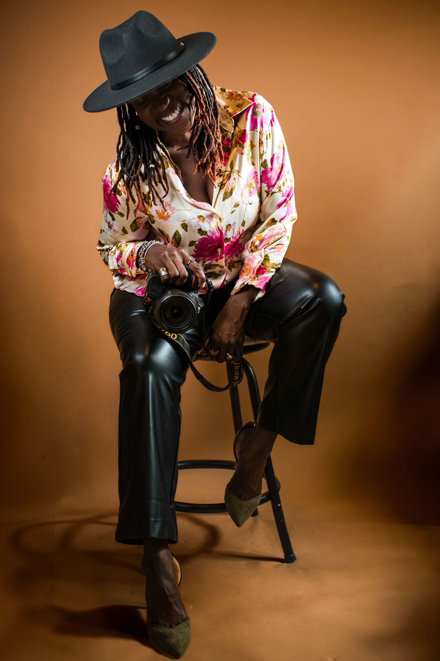 Photographer in black hat and floral blouse sits on stool holding camera against brown backdrop in studio setting.