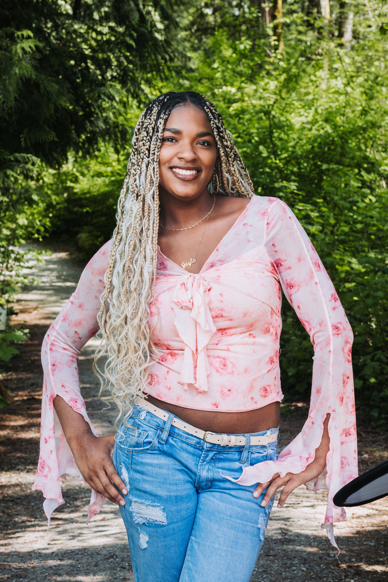 Smiling young woman with long blonde braids wearing a pink floral crop top and ripped jeans outdoors.