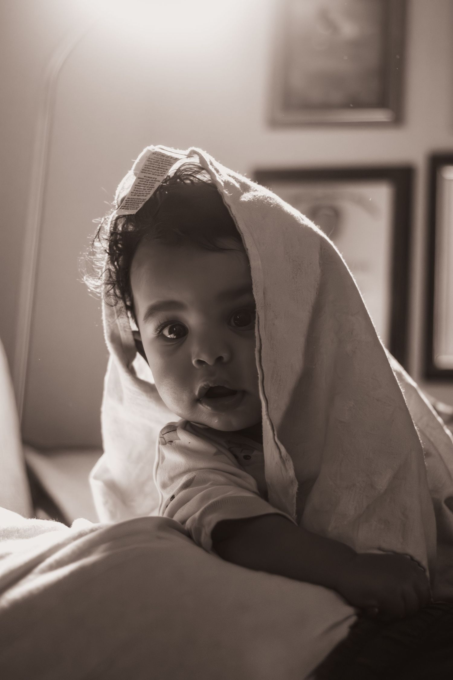 Sepia-toned photo of a cute baby peeking out from under a white blanket while sitting on a bed.