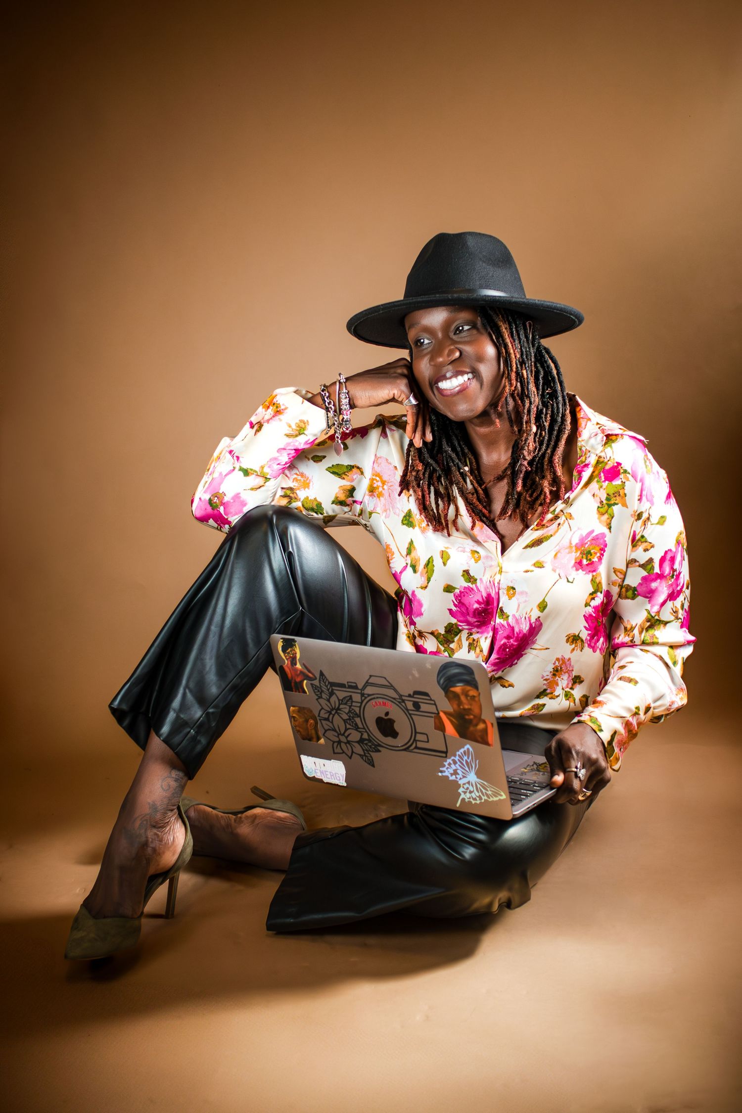 A person in a floral dress and black hat reclines while working on a laptop against a brown backdrop.