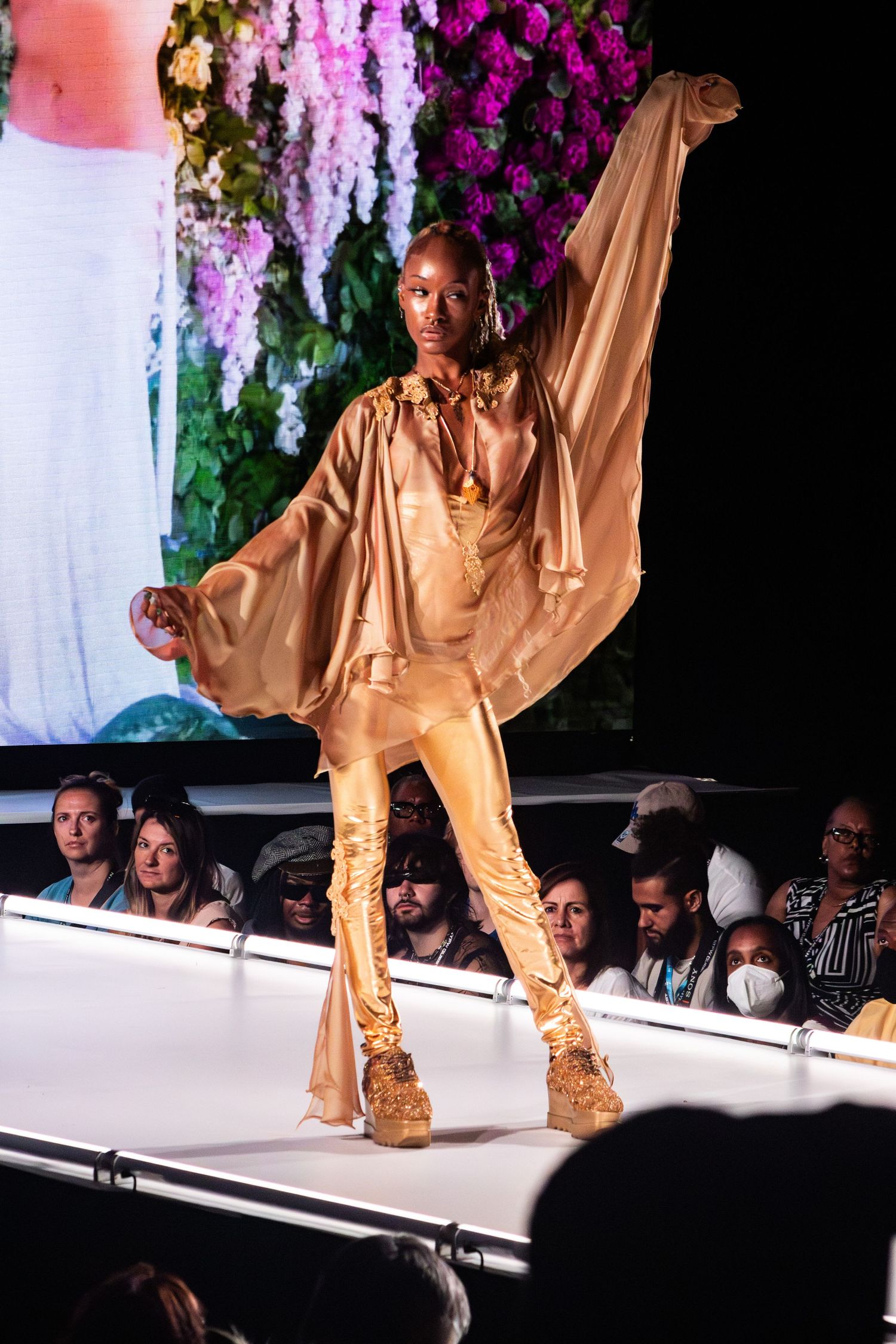 Model walks runway in flowing gold outfit against backdrop of purple flowers.
