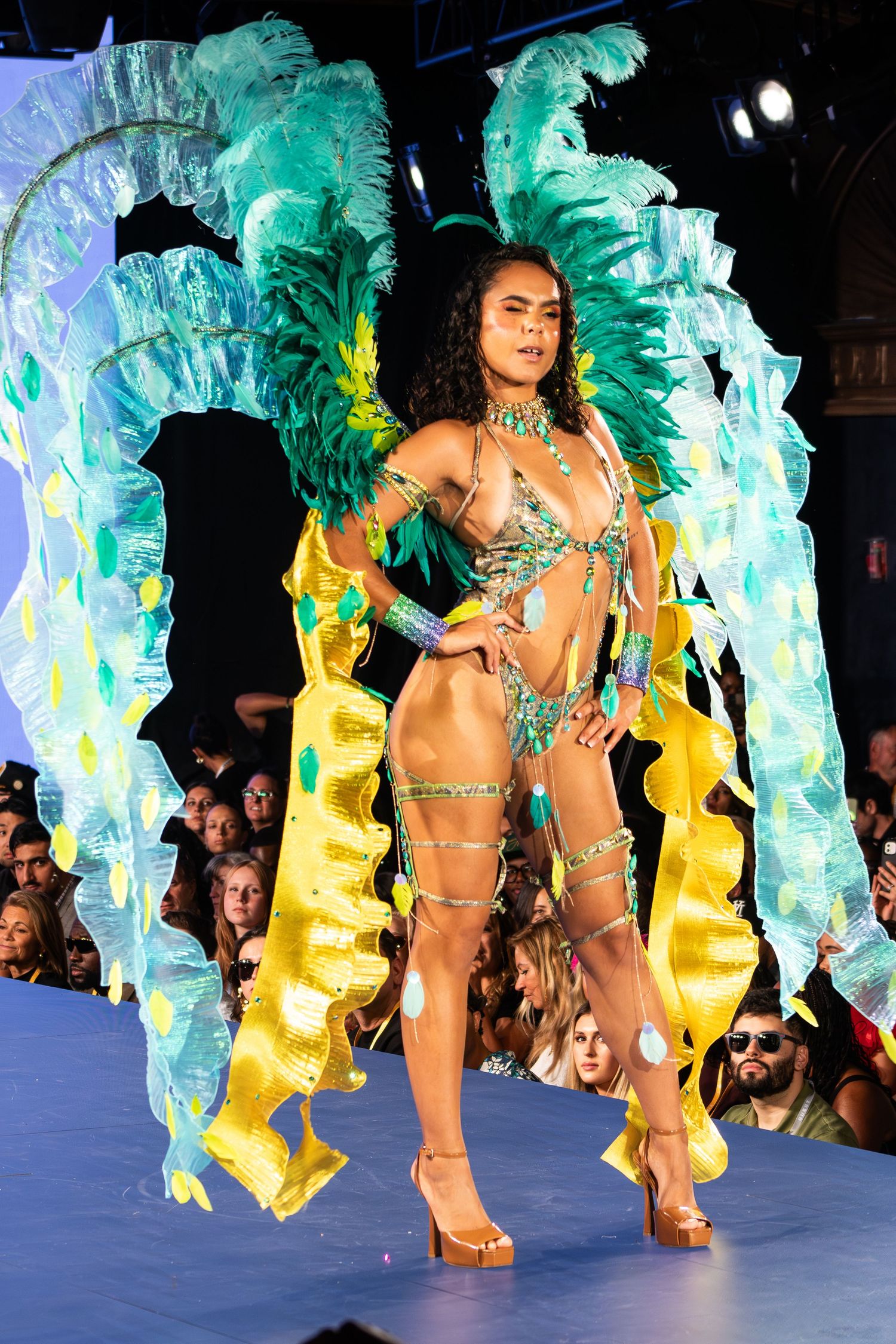 Model poses on runway in green and yellow carnival costume with large feather wings.