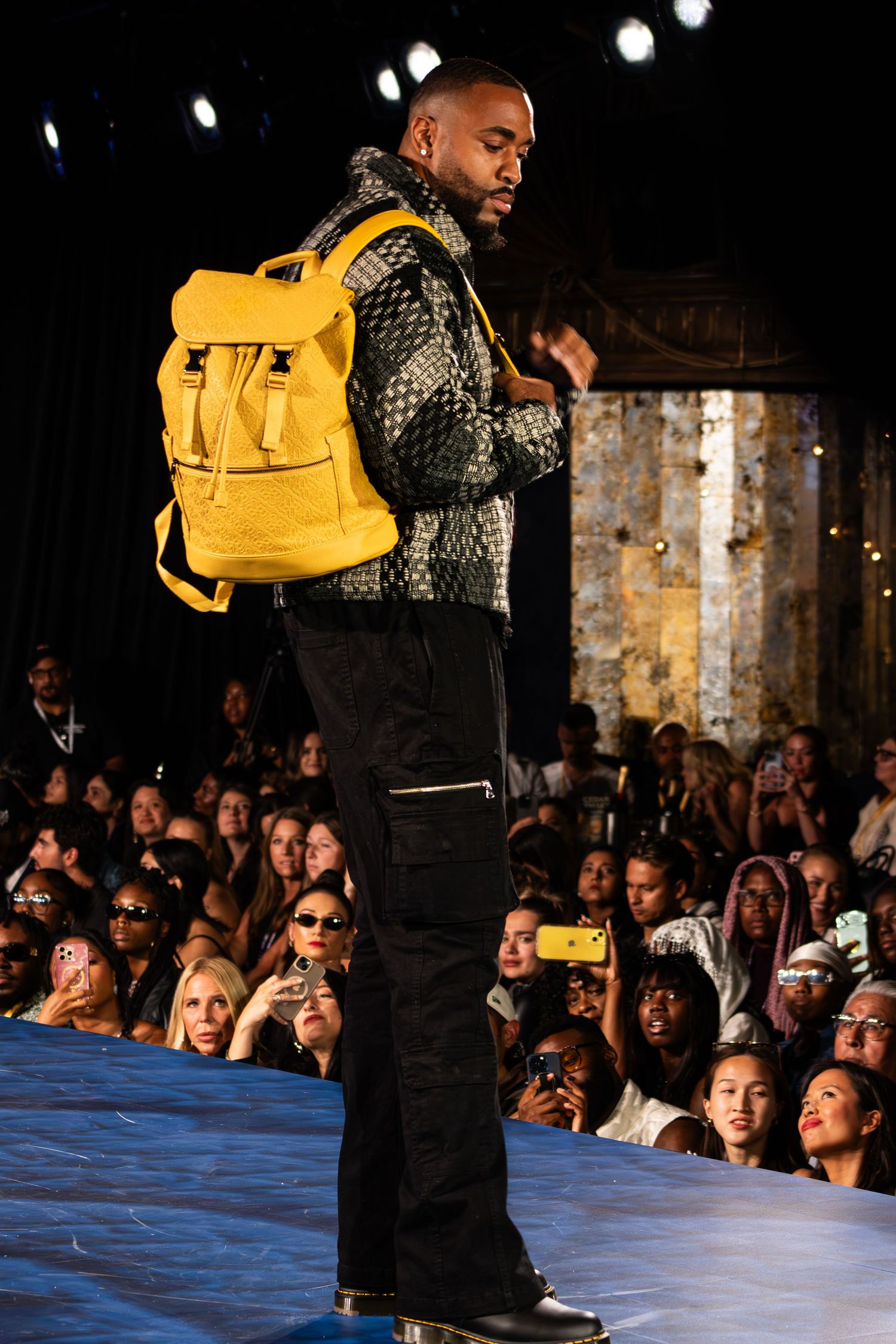 Male model on runway wearing patterned jacket with bold yellow backpack and black cargo pants at fashion show.