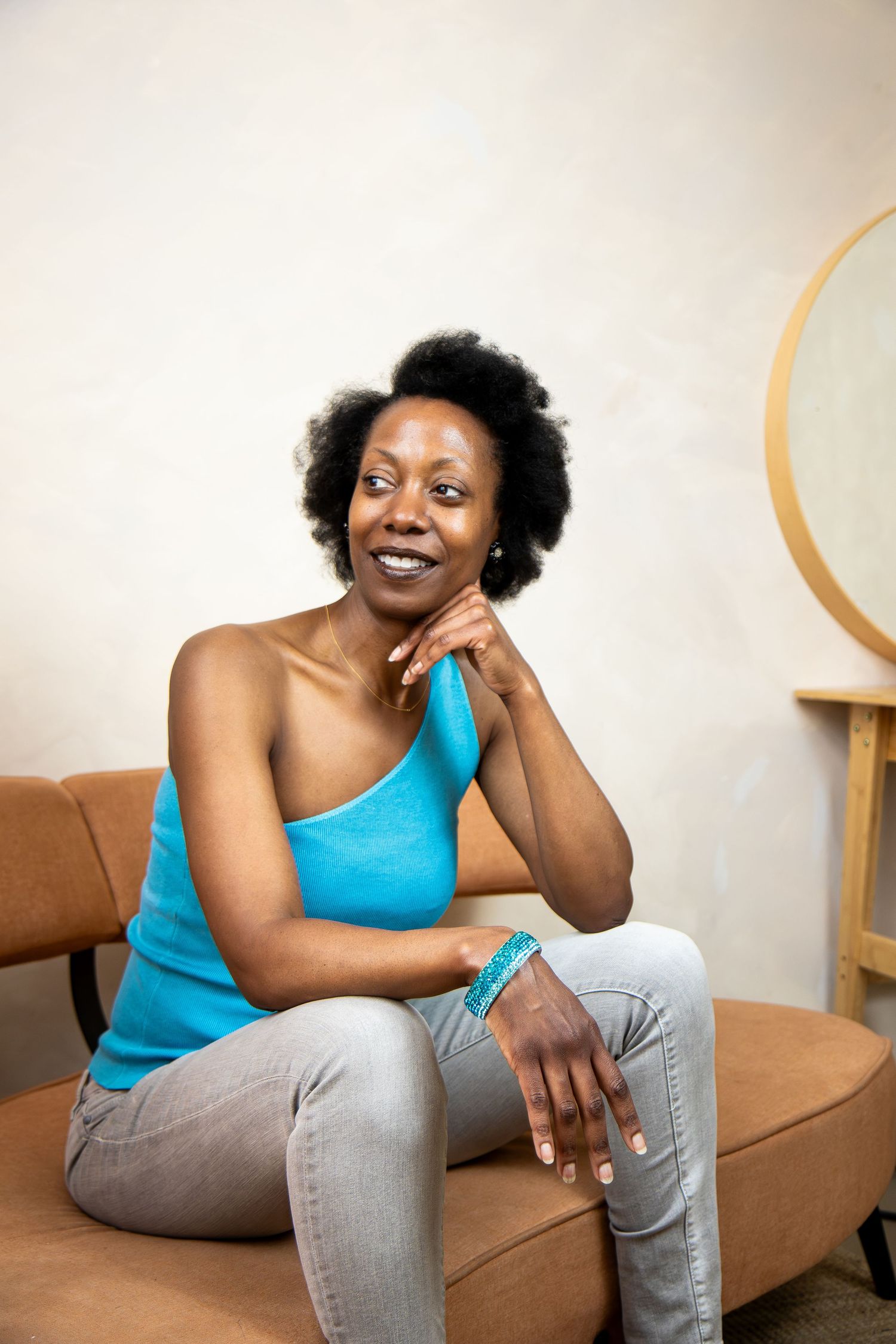 Someone in a turquoise one-shoulder top and light jeans sits casually on a brown couch with a warm smile.