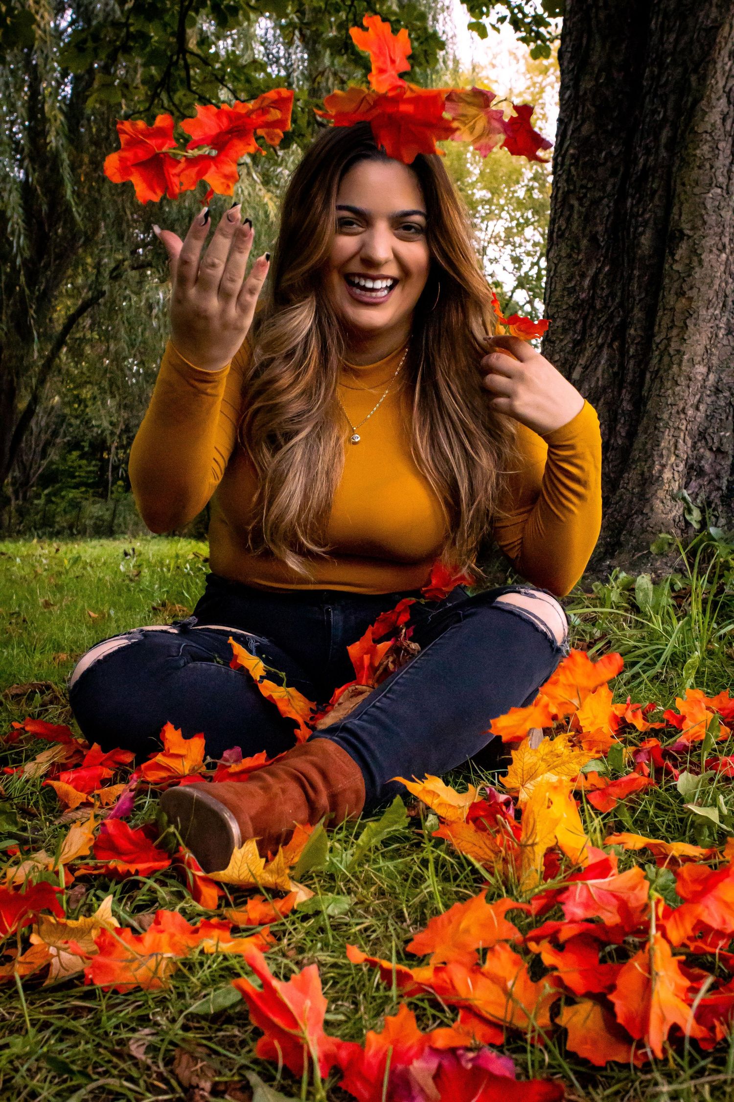 Person in yellow sweater sits among vibrant red fall leaves under a tree.