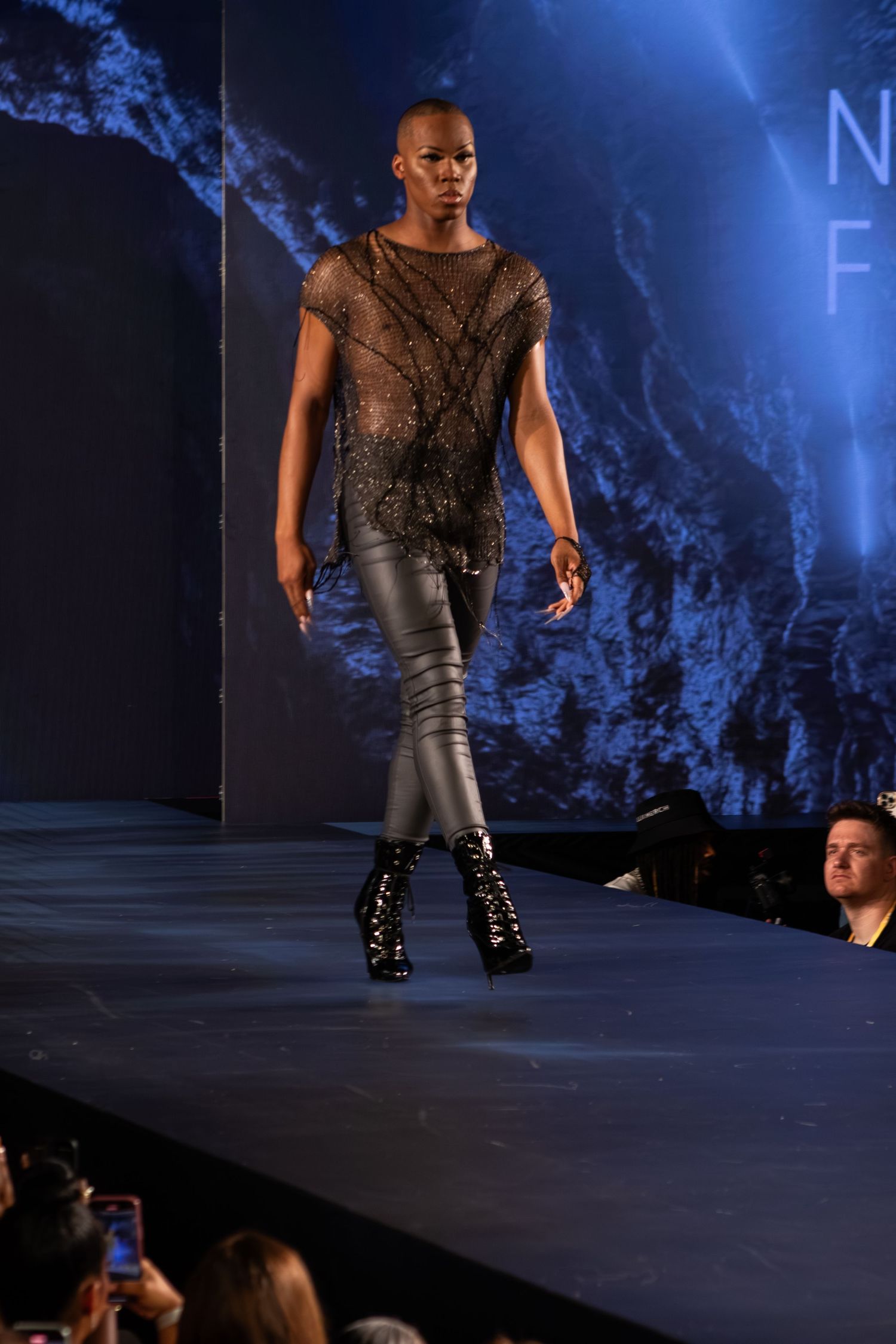 Bald model walks runway in sheer black top with metallic leggings and boots.