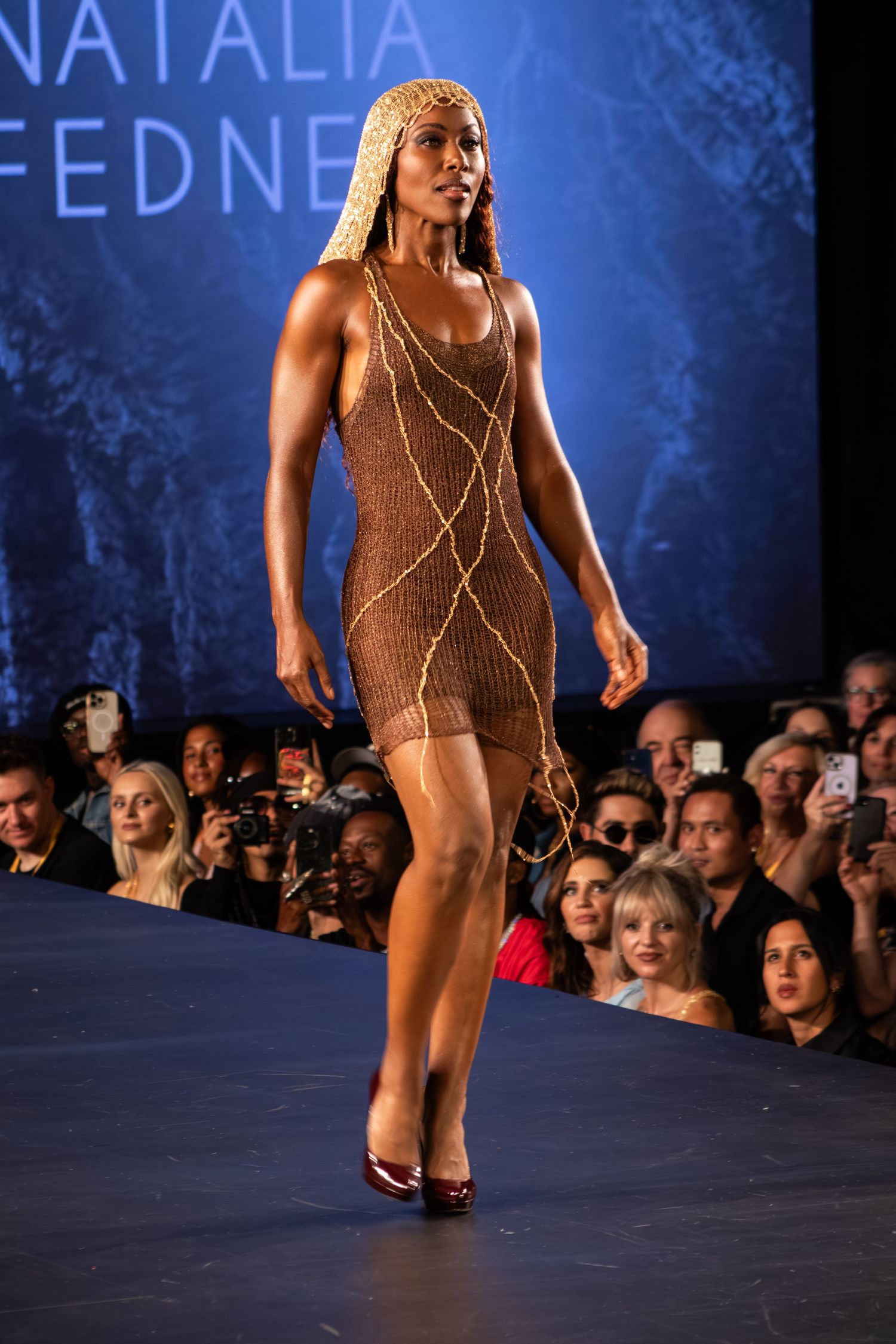 Model walks runway in brown knit dress with gold chain details at Natalia Fedner fashion show.