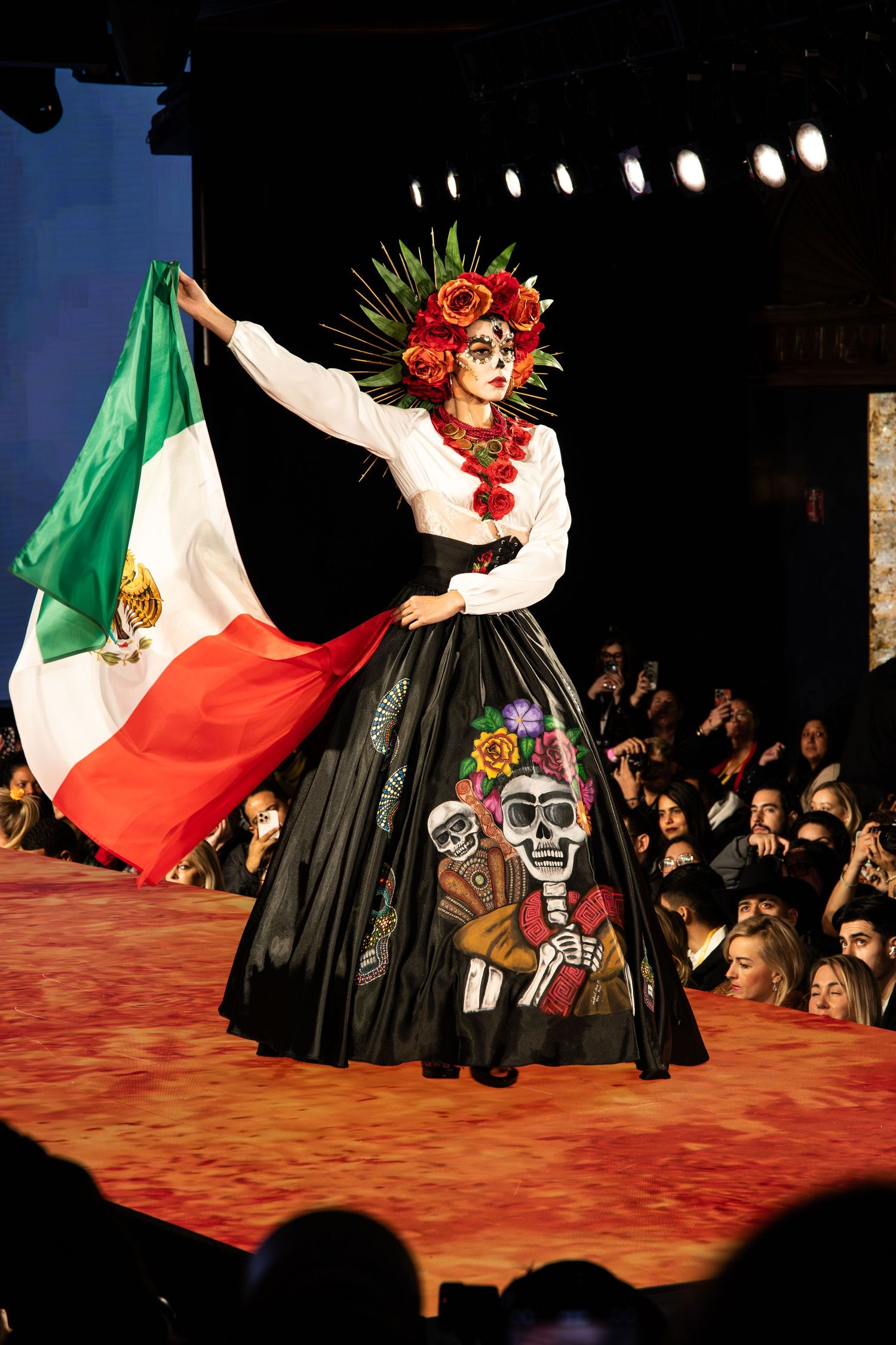 A performer in Dia de los Muertos costume with colorful skull dress and headdress waves Mexican flag on stage.