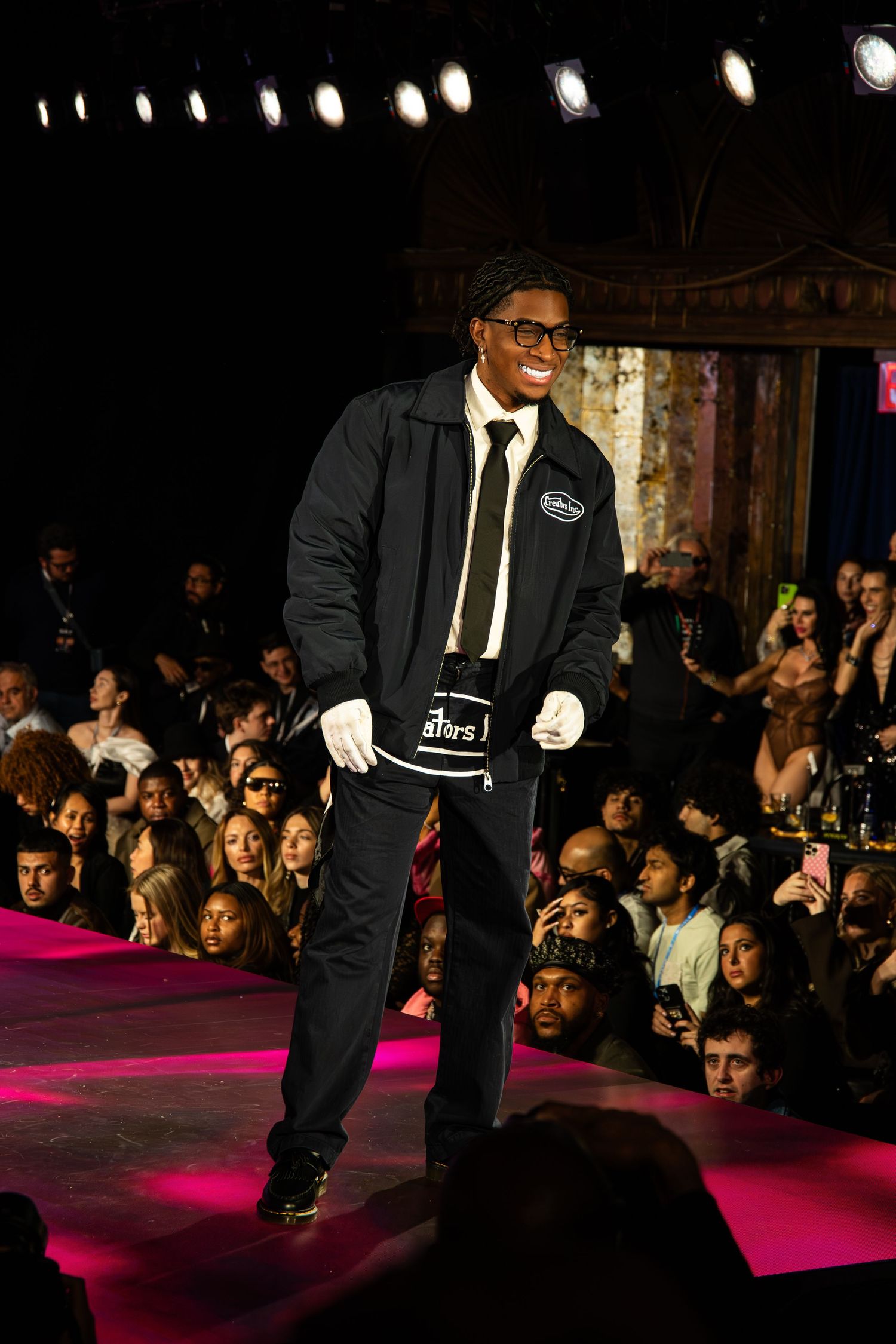 A performer wearing a black suit and tie walks the runway under stage lights during a fashion show with spectators watching.