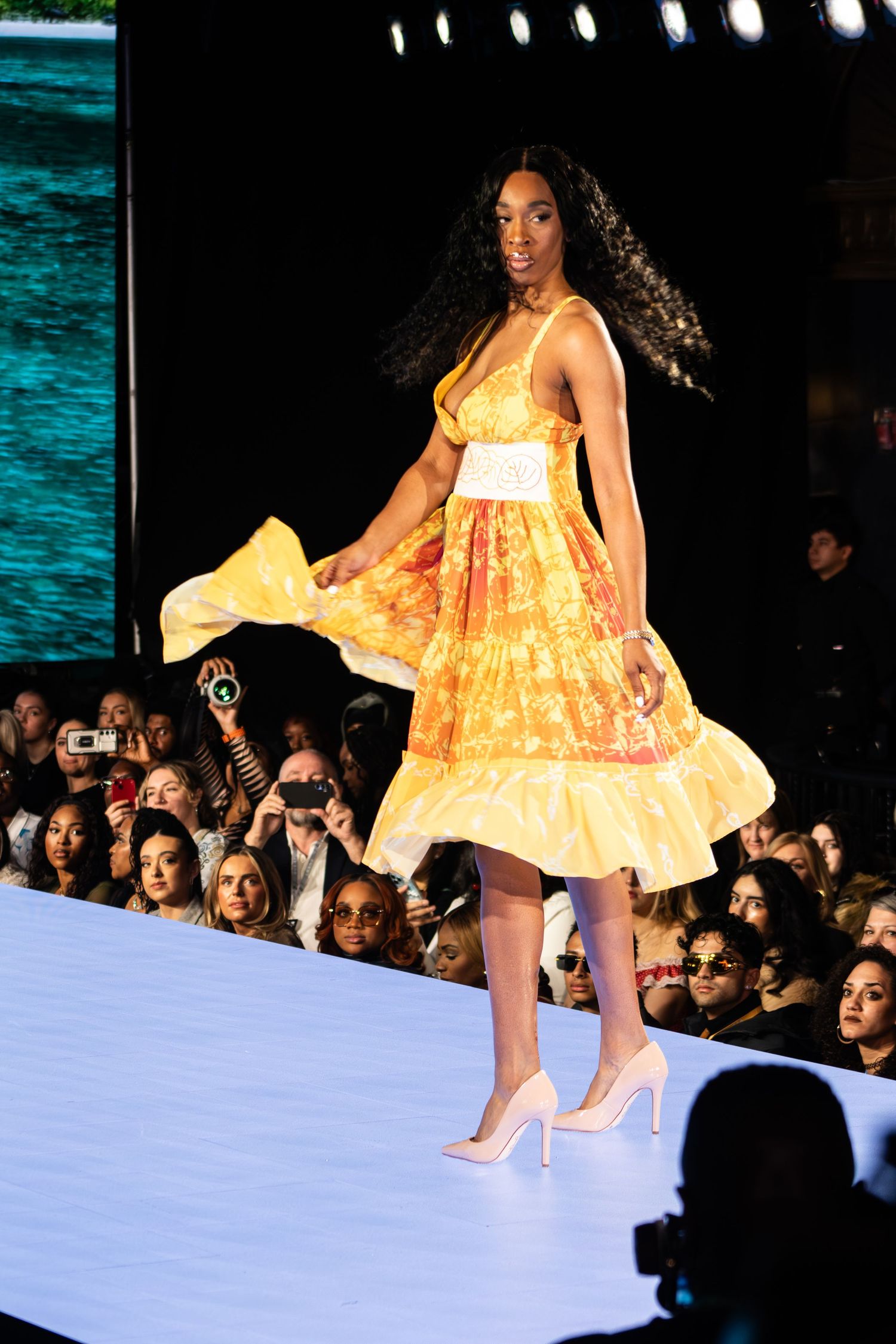 Model walks runway in flowing yellow dress with white waistband and nude heels at fashion show.