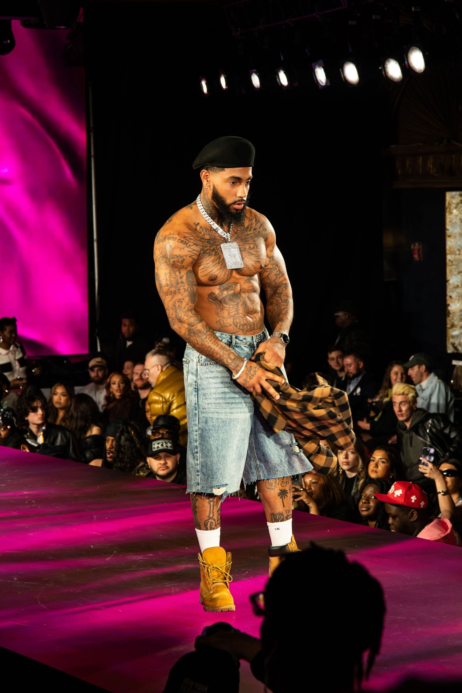 A muscular model in denim shorts and boots walks a pink-lit fashion runway while audience members watch in silhouette.