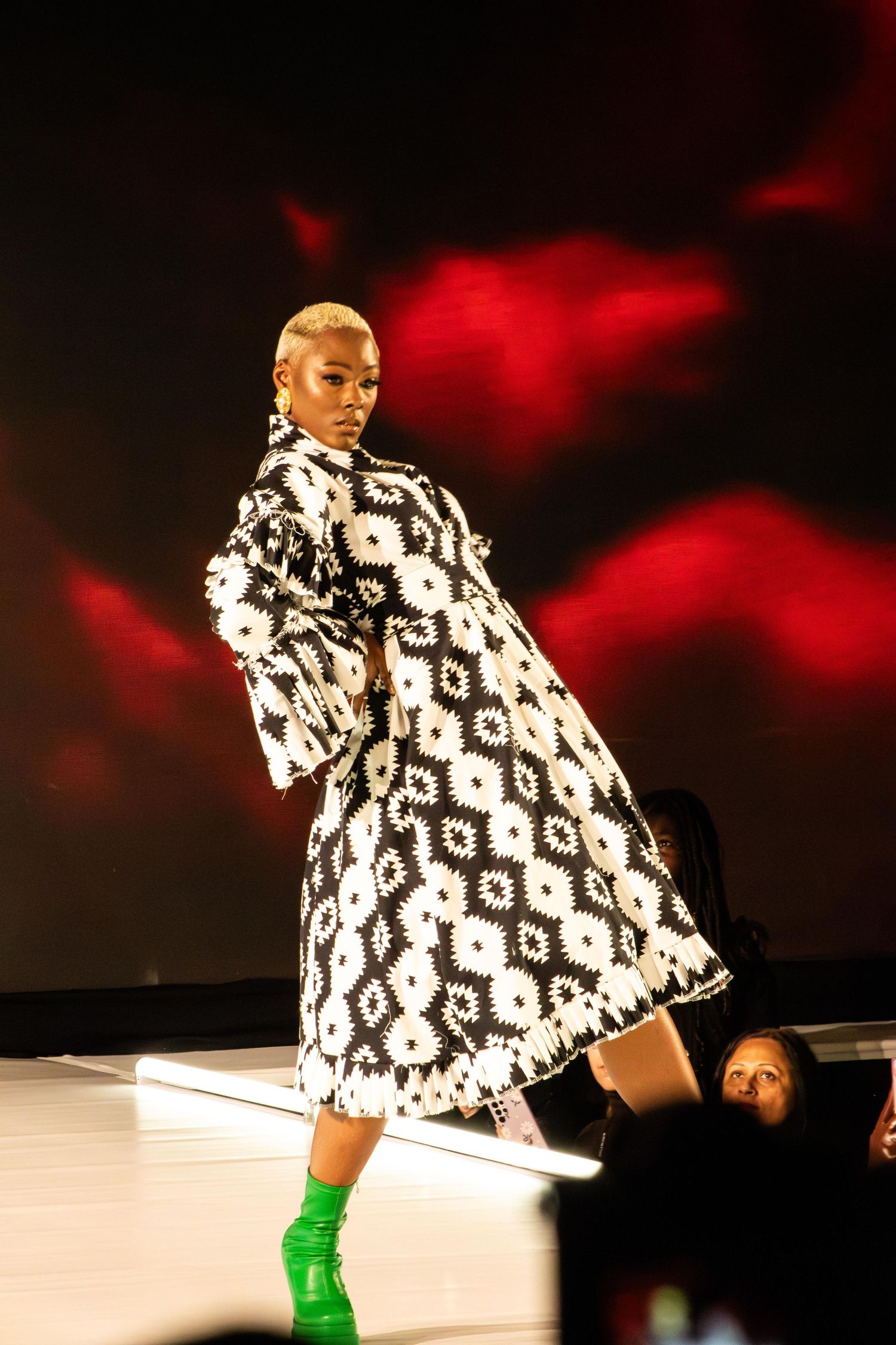 Dramatic black and white patterned coat with green boots on red-lit runway.