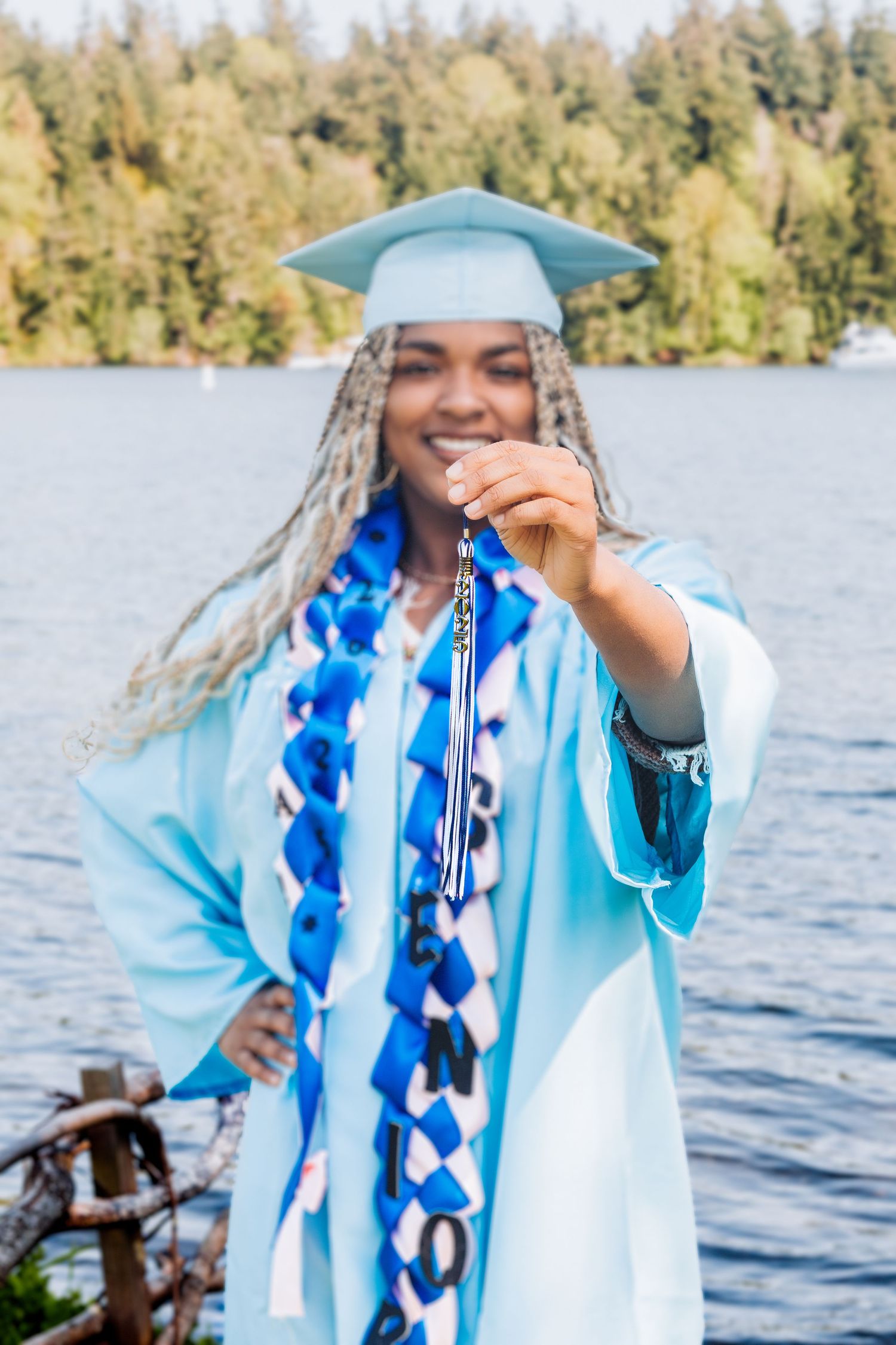 A smiling graduate in a light blue cap and gown holds up a tassel by a lake with trees in the background.